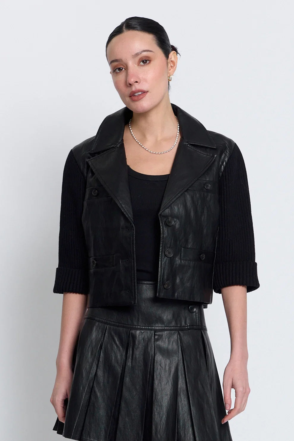 A woman modeling a black faux-leather cropped jacket vest over a black ribbed top and a black faux-leather pleated skirt, against a light gray studio background.