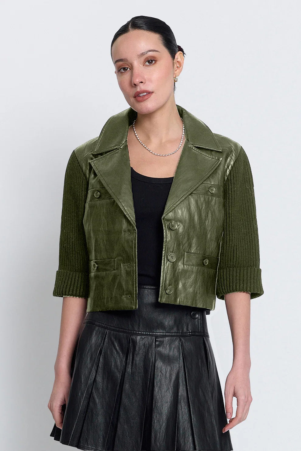 A woman modeling a cropped olive green jacket with a mix of textured ribbed sleeves and smooth front panels, worn over a black top and black pleated skirt against a light gray studio background.