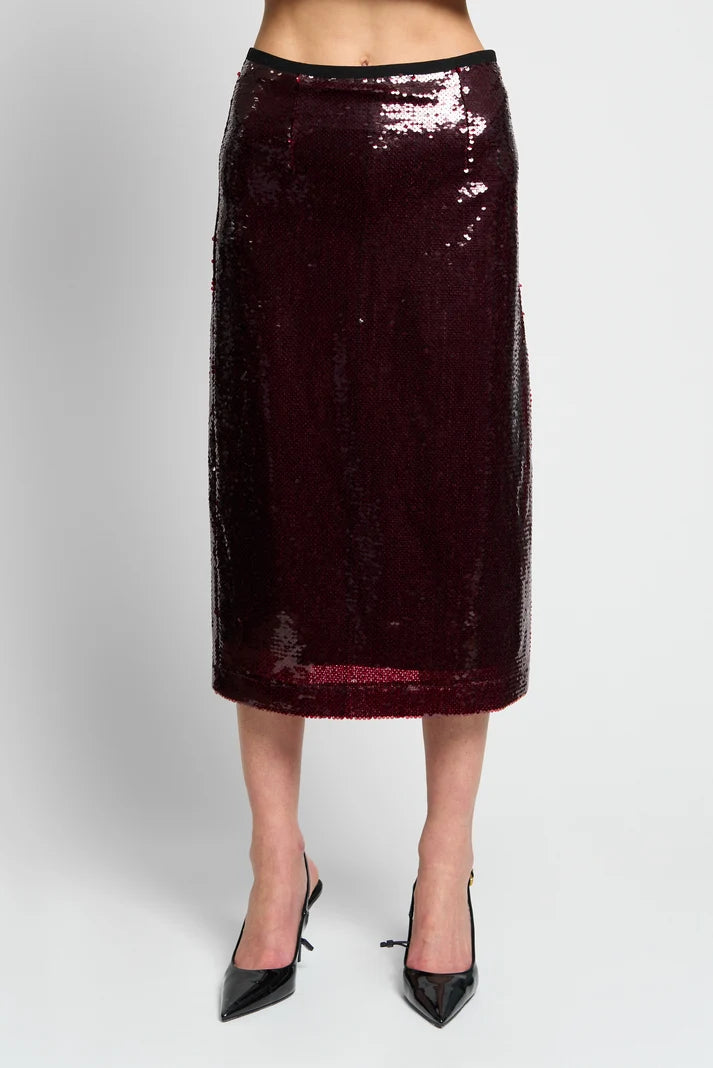 A dark burgundy sequin midi skirt worn by a model, paired with black pointed-toe heels, shown against a light gray studio background.