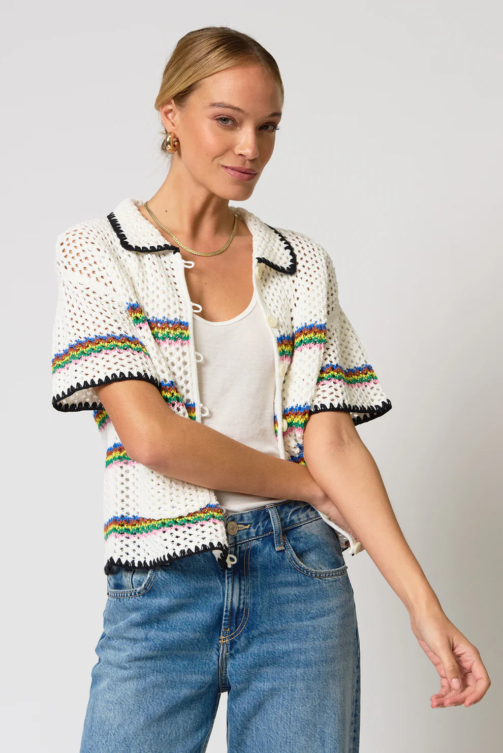 Woman wearing a white crochet cardigan with rainbow accents and blue jeans on a plain background