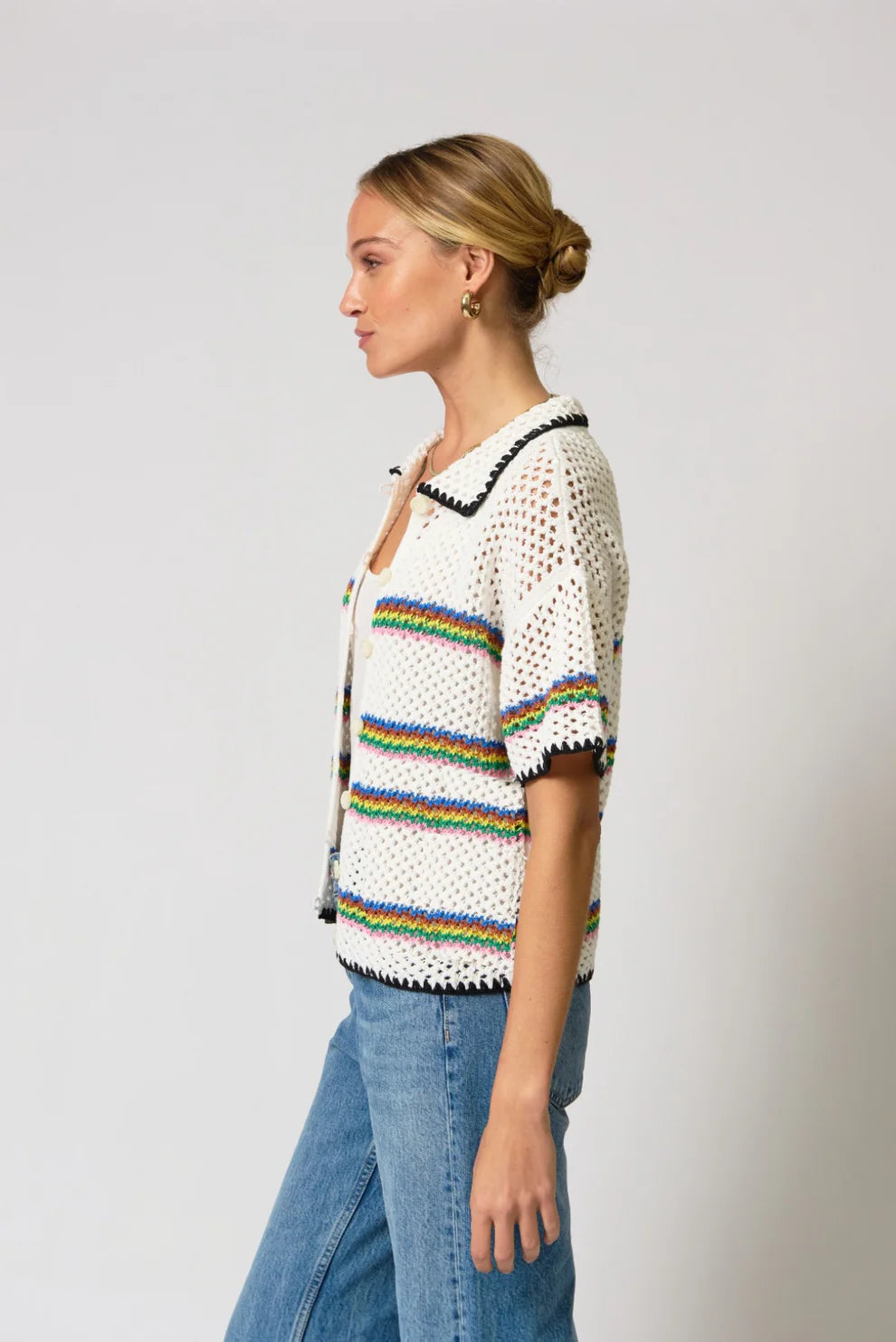 Woman wearing a white crochet top with rainbow stripes and blue jeans on a light gray background