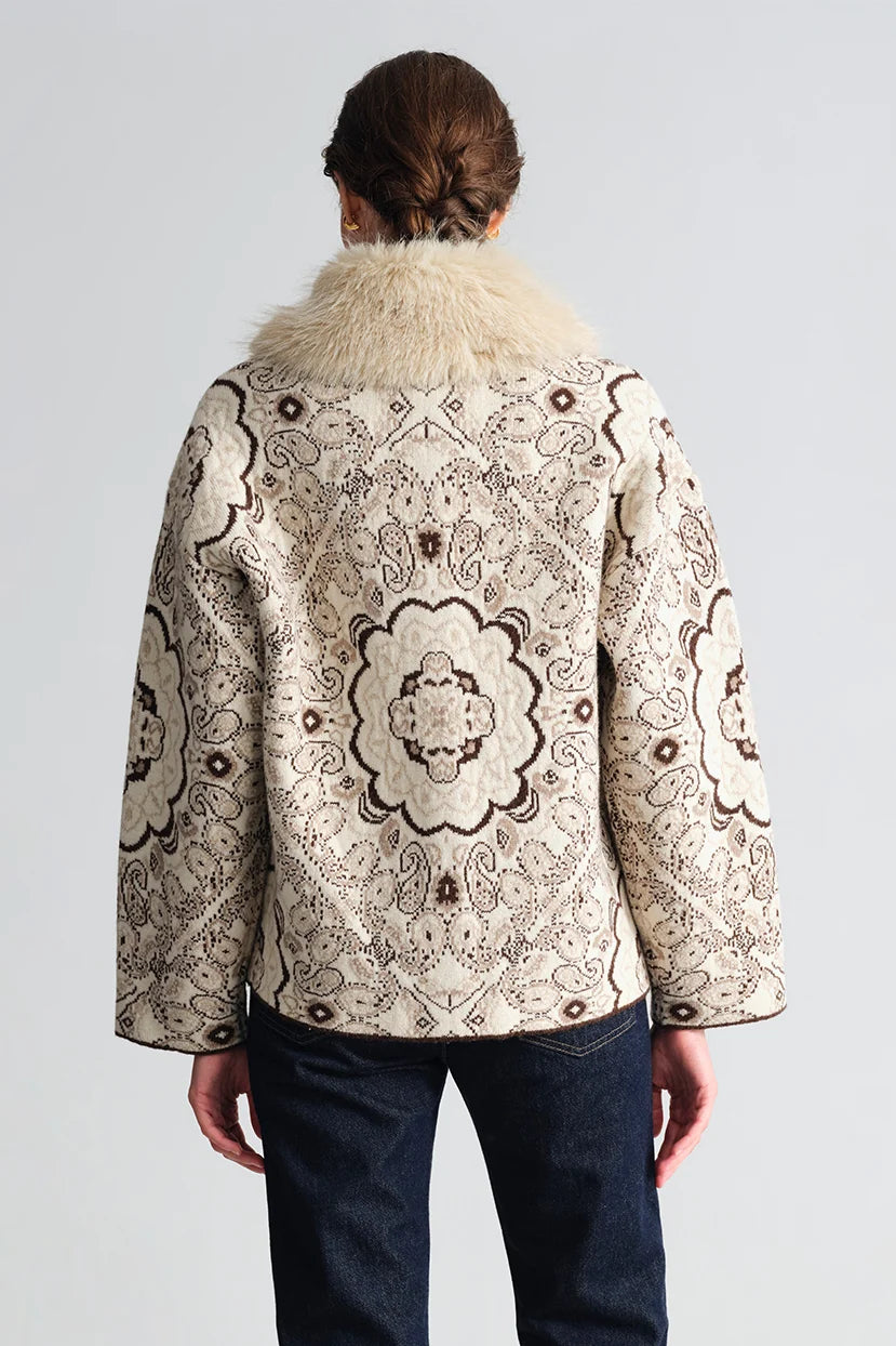 A model viewed from the back wearing a cropped jacket with an intricate beige and brown floral/medallion pattern and a fluffy cream faux-fur collar, paired with dark blue jeans against a light gray studio background.