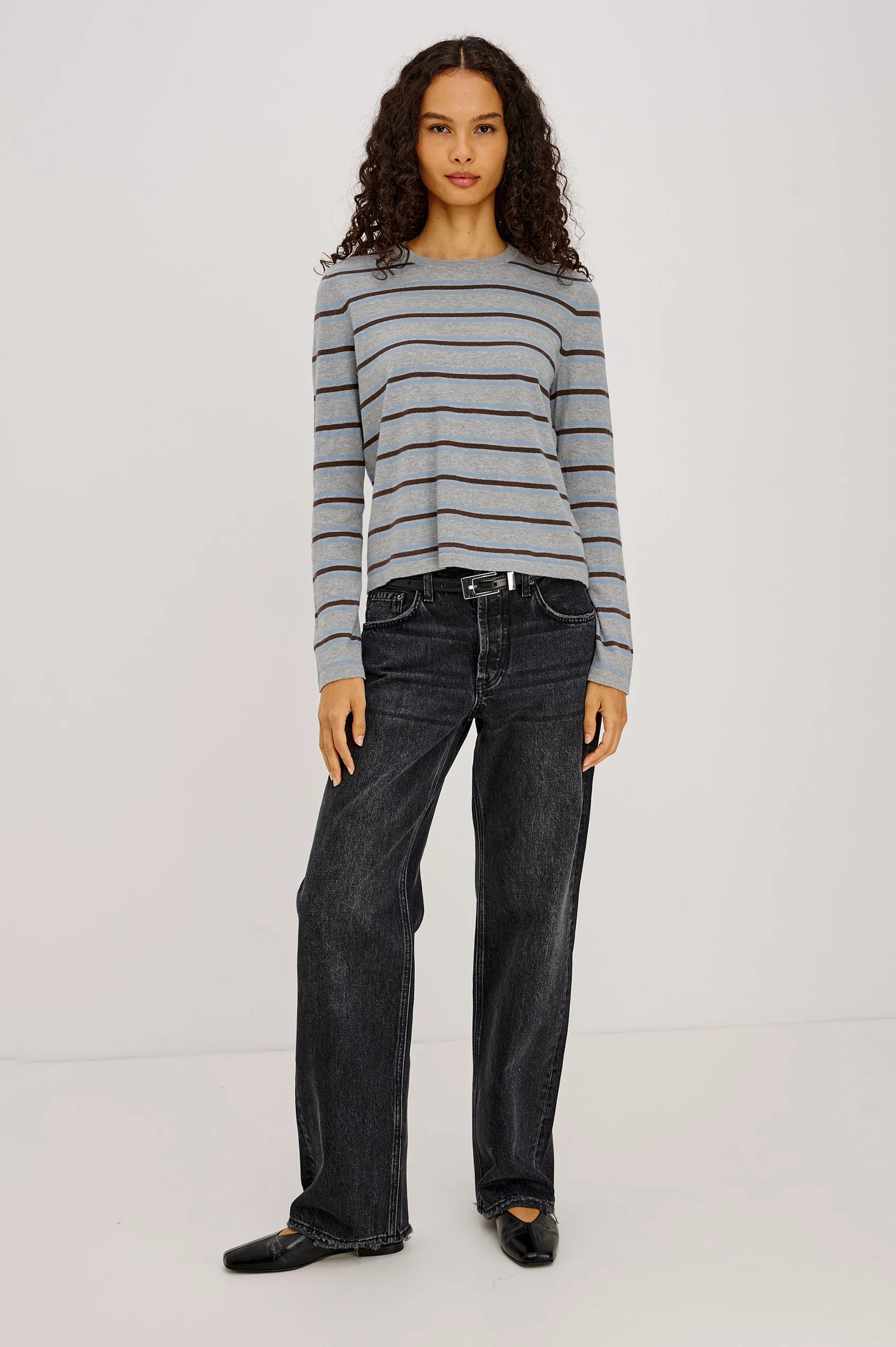 Woman wearing a gray and blue striped sweater with black jeans and a ballet flat on a plain background