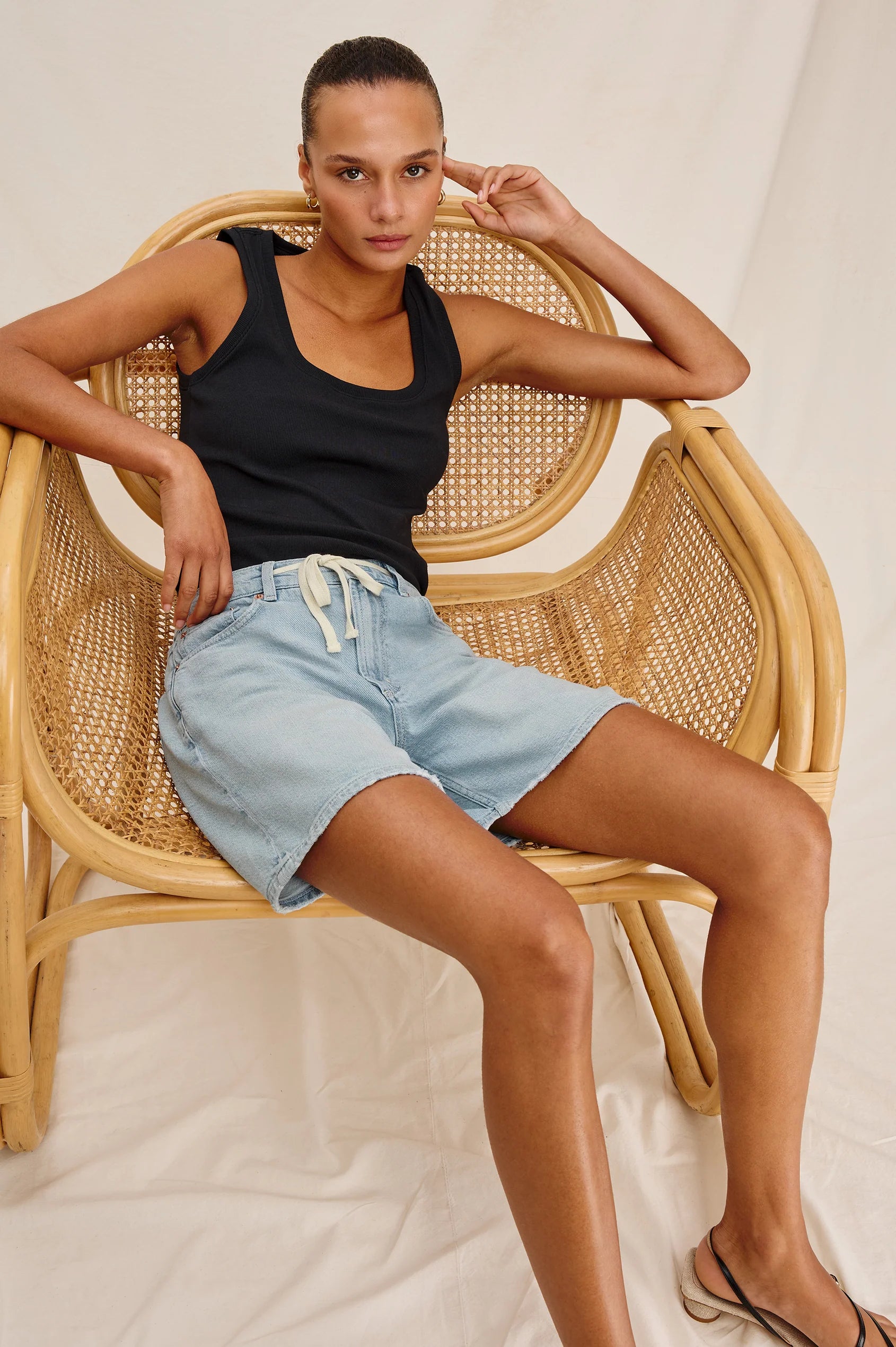 Woman sitting on a wicker chair wearing a black tank top and light blue denim shorts.