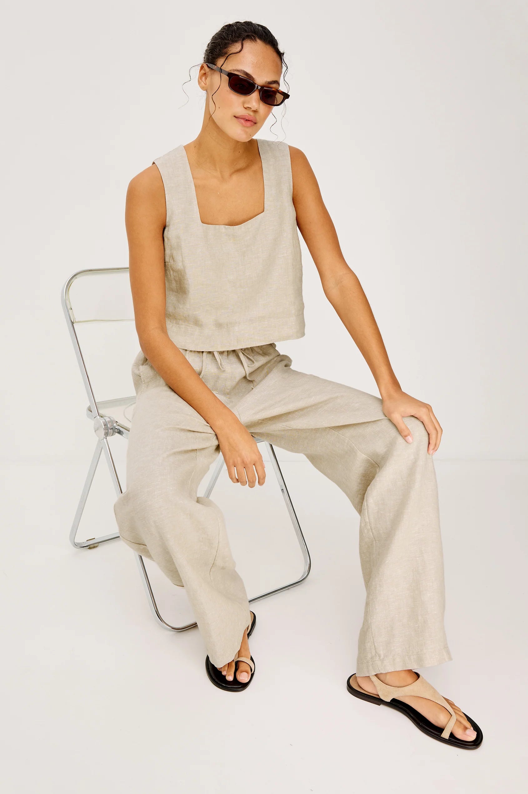 Woman sitting on a chair wearing a beige sleeveless top and pants with sunglasses on a white background