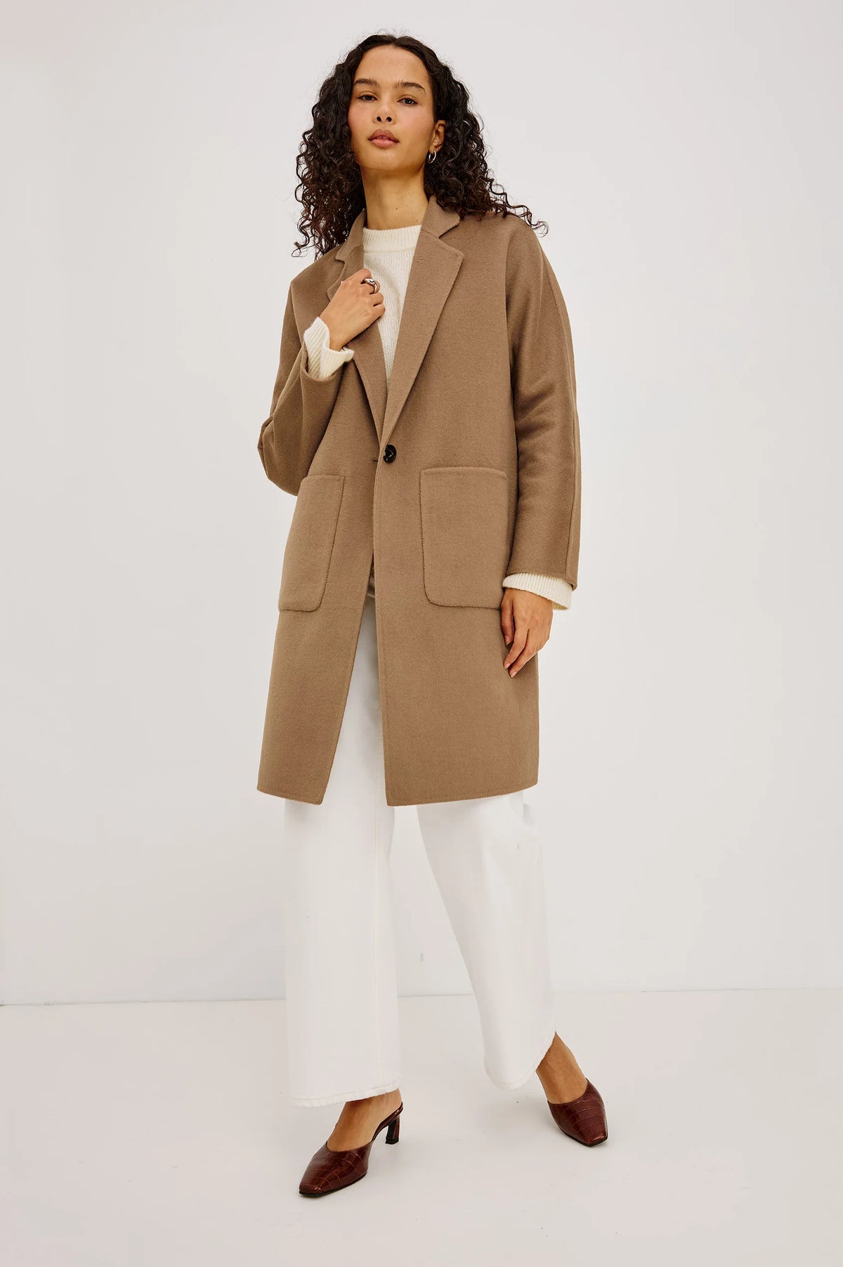 A woman modeling a camel brown wool-blend coat with a single-breasted button closure and large front patch pockets, worn over a cream top and white wide-leg trousers, against a plain light gray studio background.