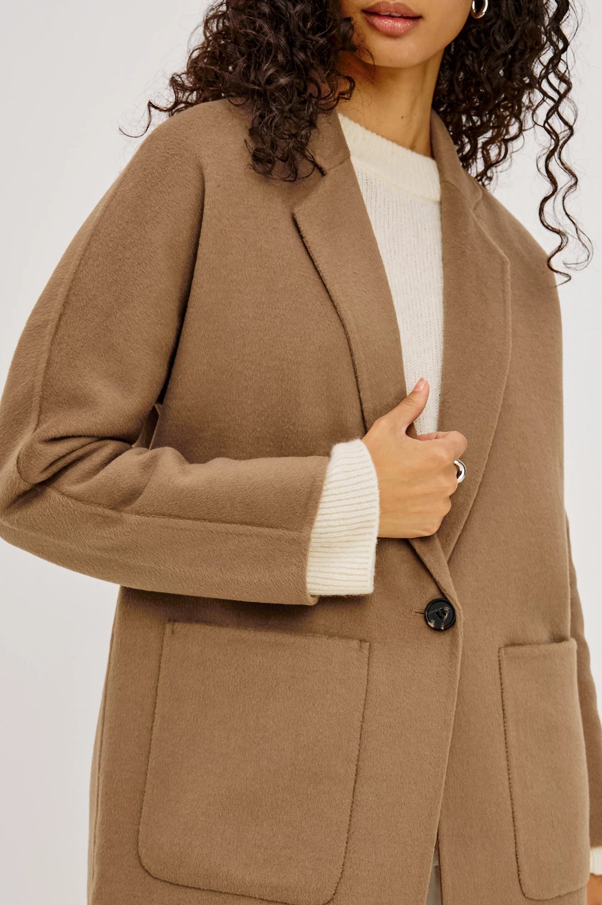 A close-up of a woman wearing a camel brown oversized blazer over a cream ribbed sweater, with her hand resting on the lapel.