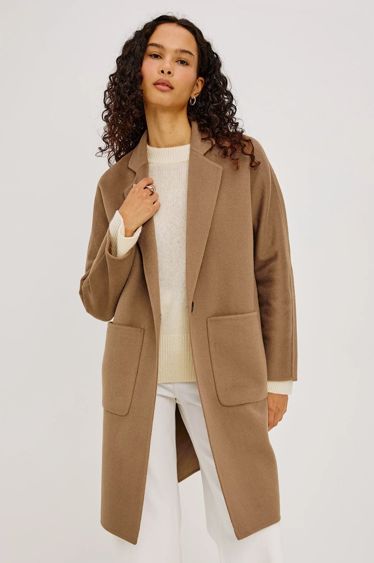 A woman with curly dark hair models a mid-length camel brown coat over a cream knit top and white pants, photographed against a plain light gray studio background.