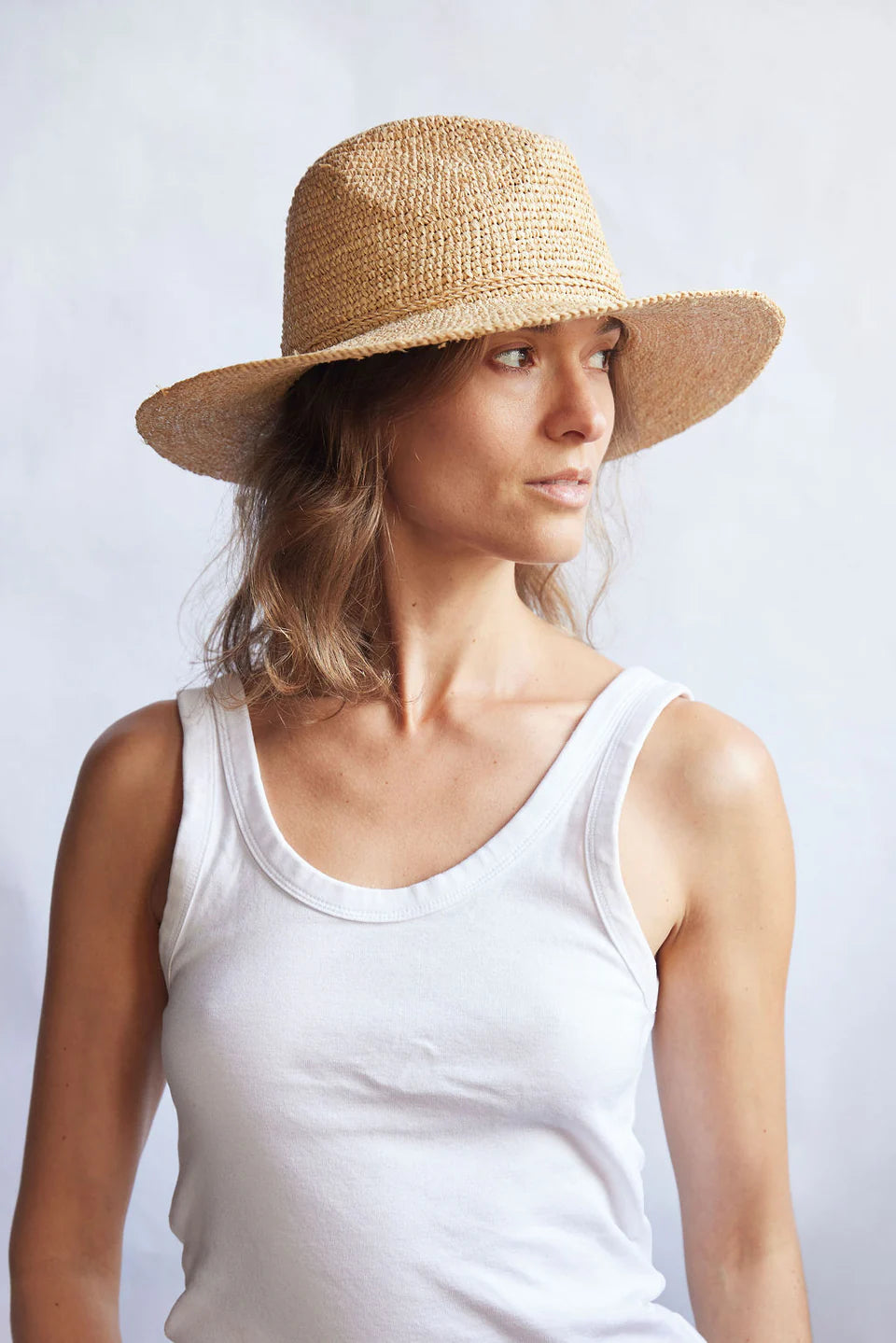 Woman wearing a straw hat and white tank top against a plain background