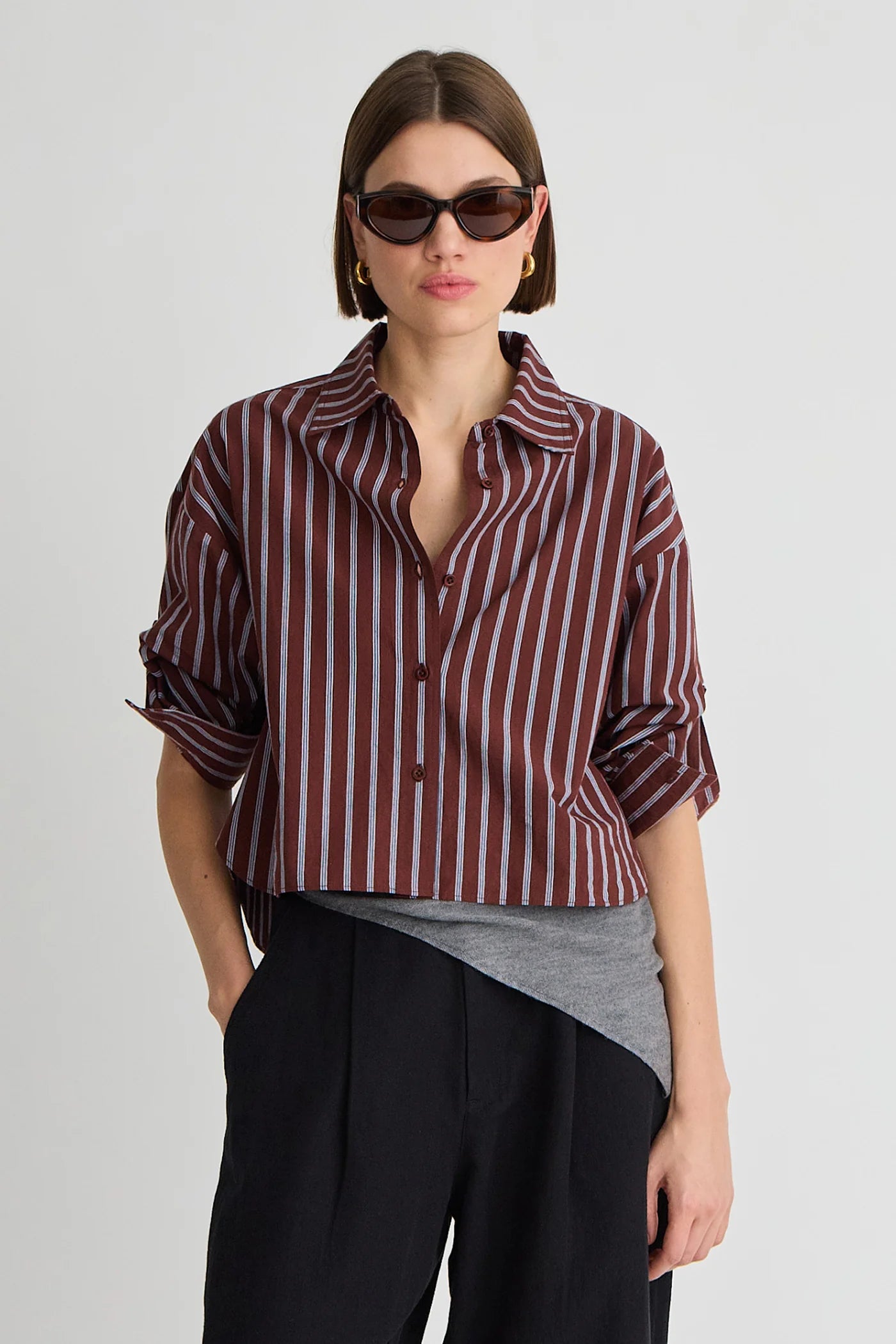 A model wearing a cropped button-up shirt with vertical burgundy and white stripes, styled with a grey undershirt, black trousers, and dark sunglasses against a light gray background.