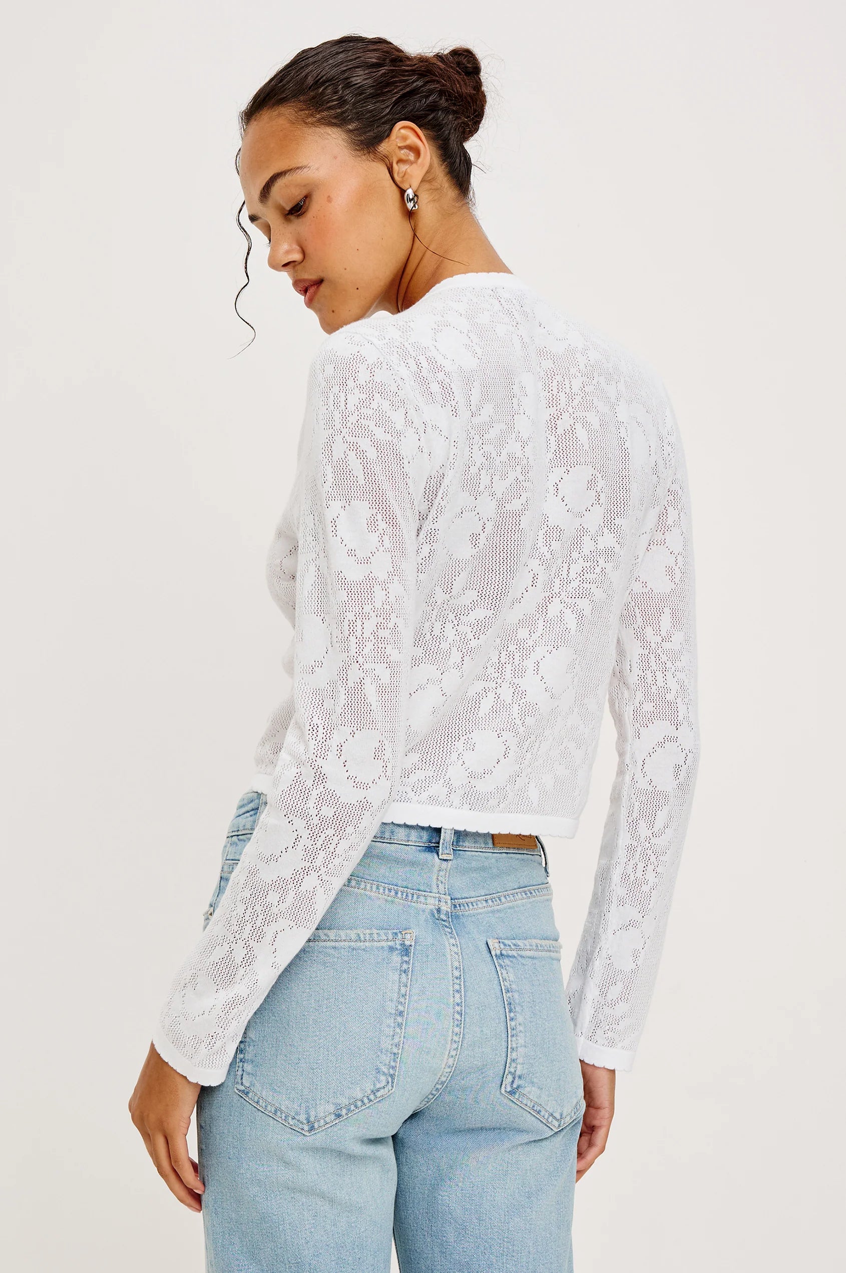 Woman wearing a white lace long-sleeve top and light blue jeans on a plain background