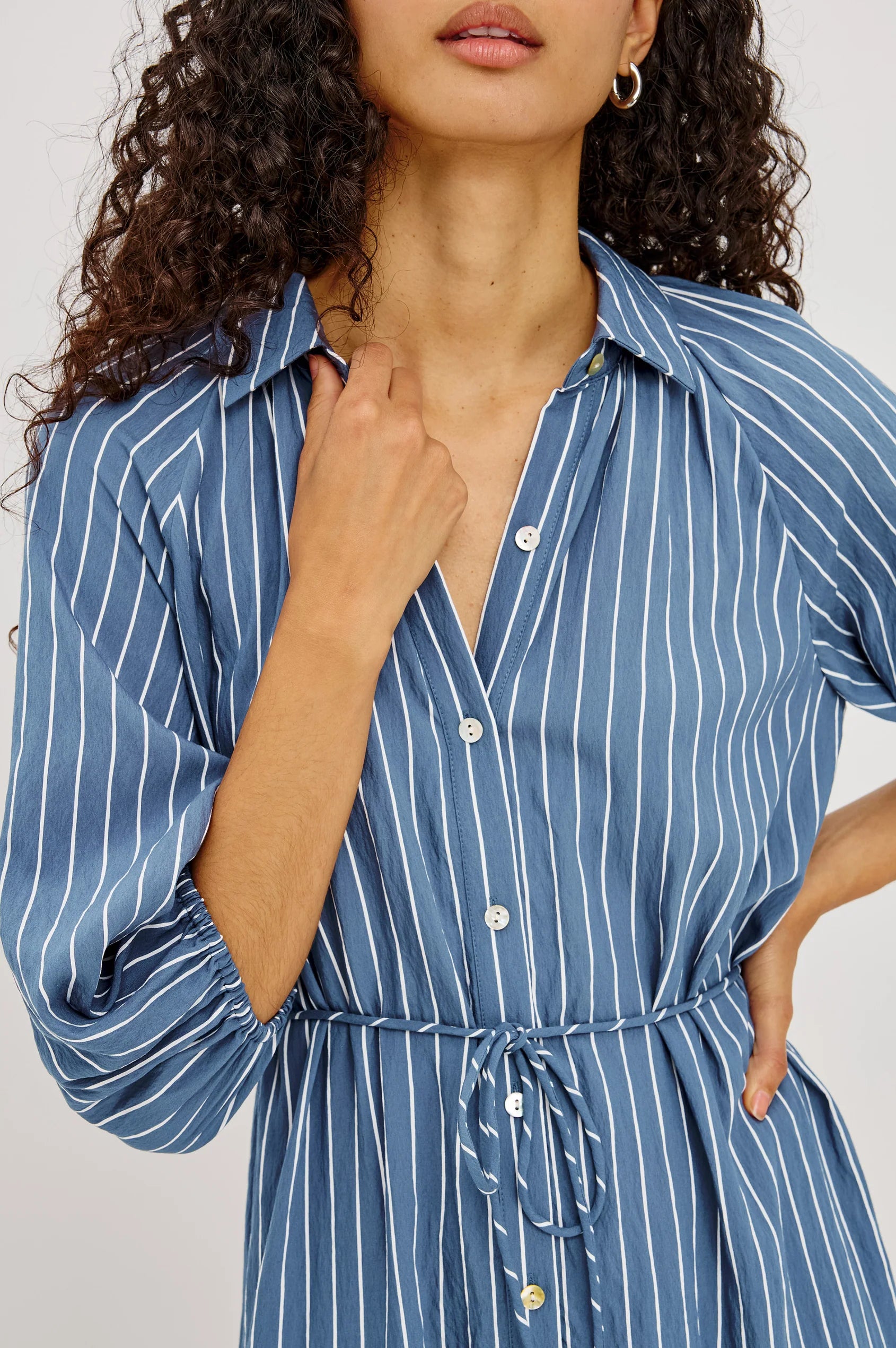 A woman modeling a blue and white vertically striped midi dress with puff sleeves and a tiered skirt, shown from a close up front angle from the mouth to the waist against a plain white studio background.