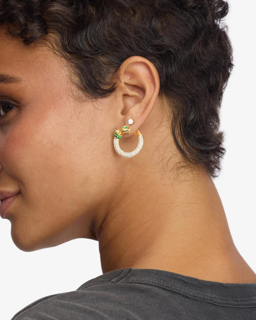 Close-up of a person wearing gold hoop earrings with green gemstones on a neutral background