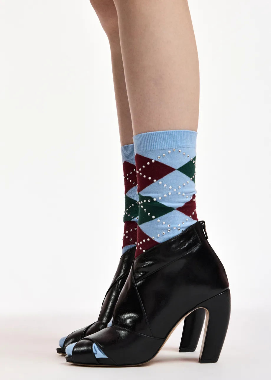 A close-up of a person's legs wearing patterned crew socks with black peep-toe high-heeled ankle boots against a plain white studio background.