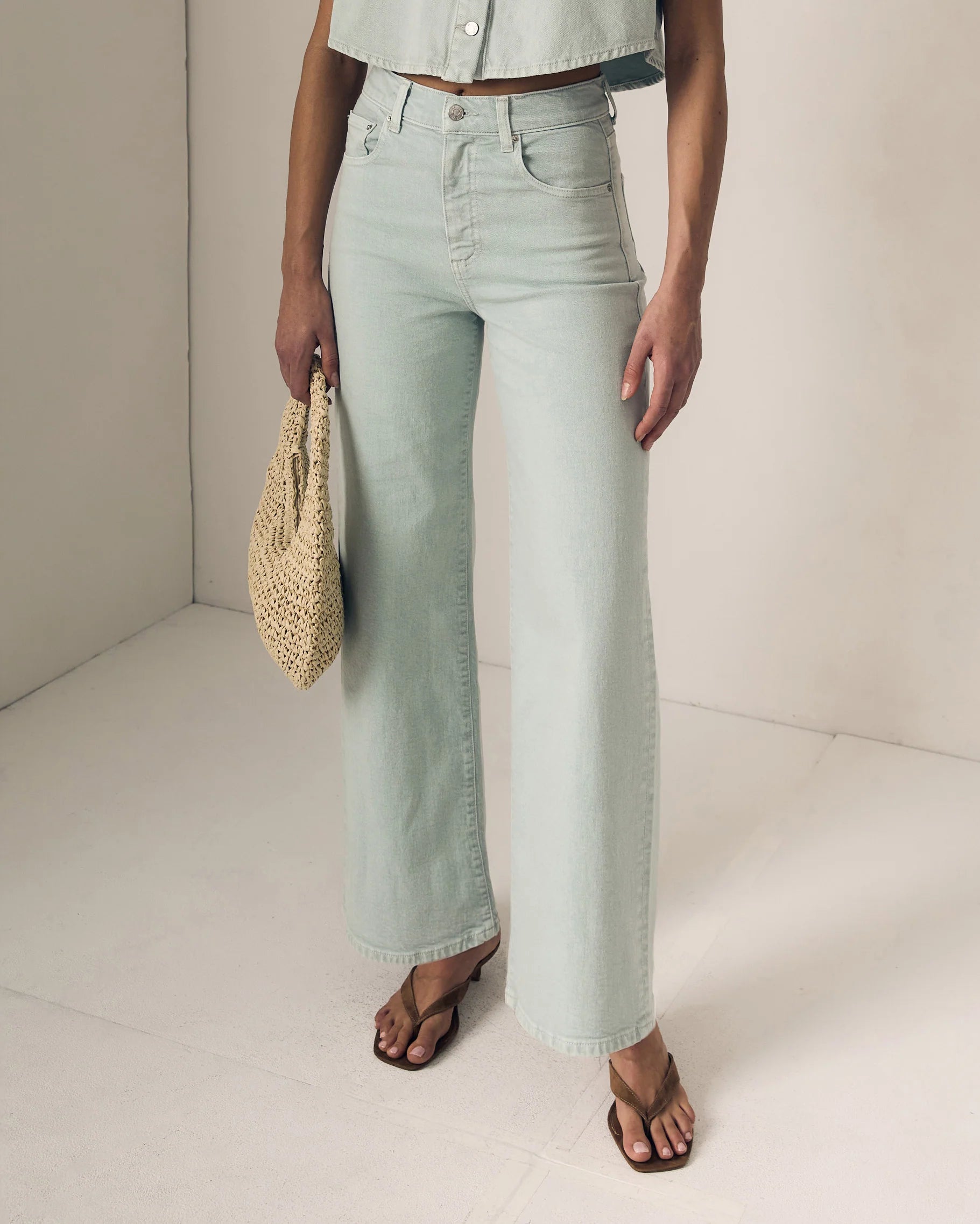 Person wearing light blue jeans holding a woven bag on a plain background