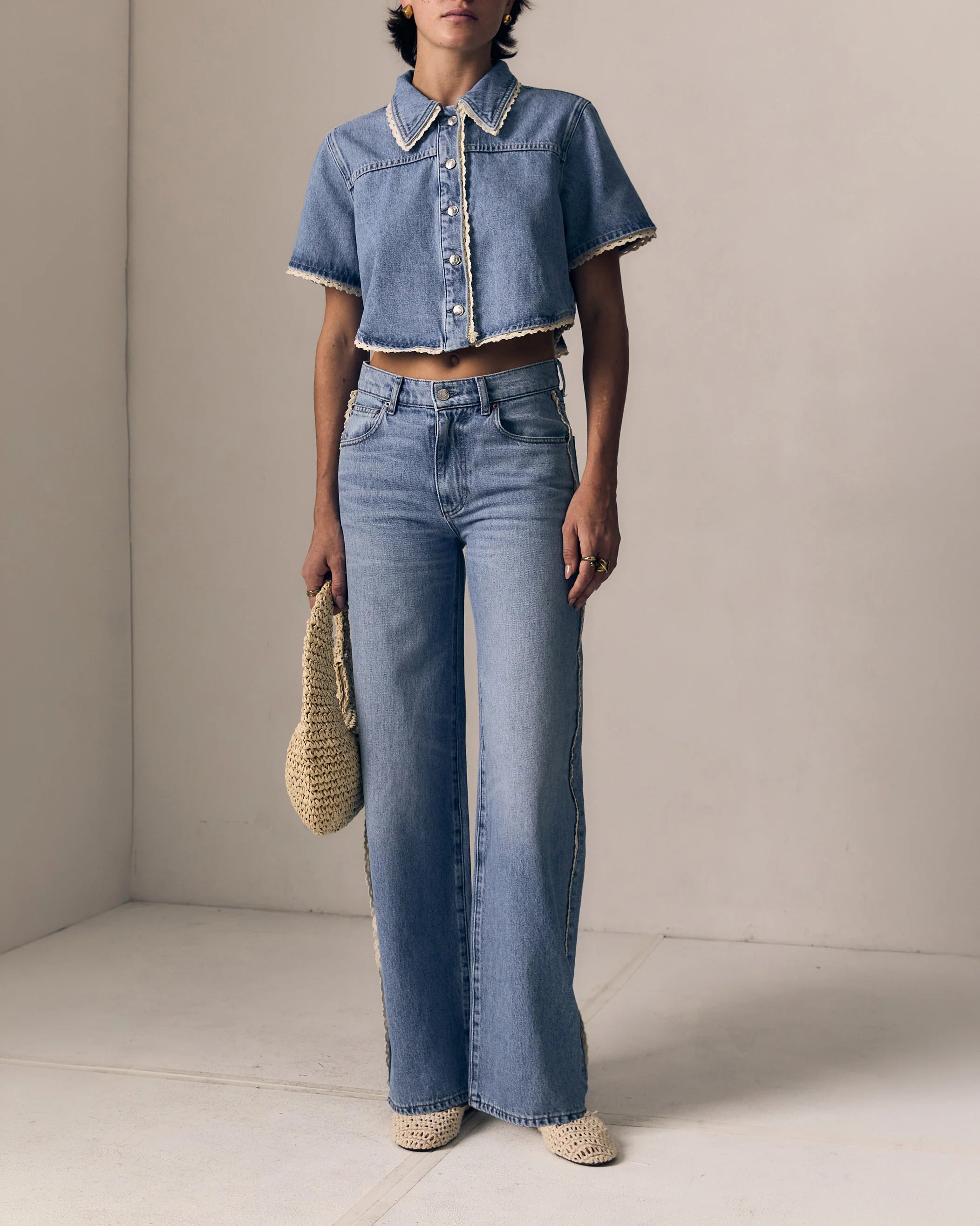 Person wearing a denim jacket and jeans holding a woven bag against a neutral background