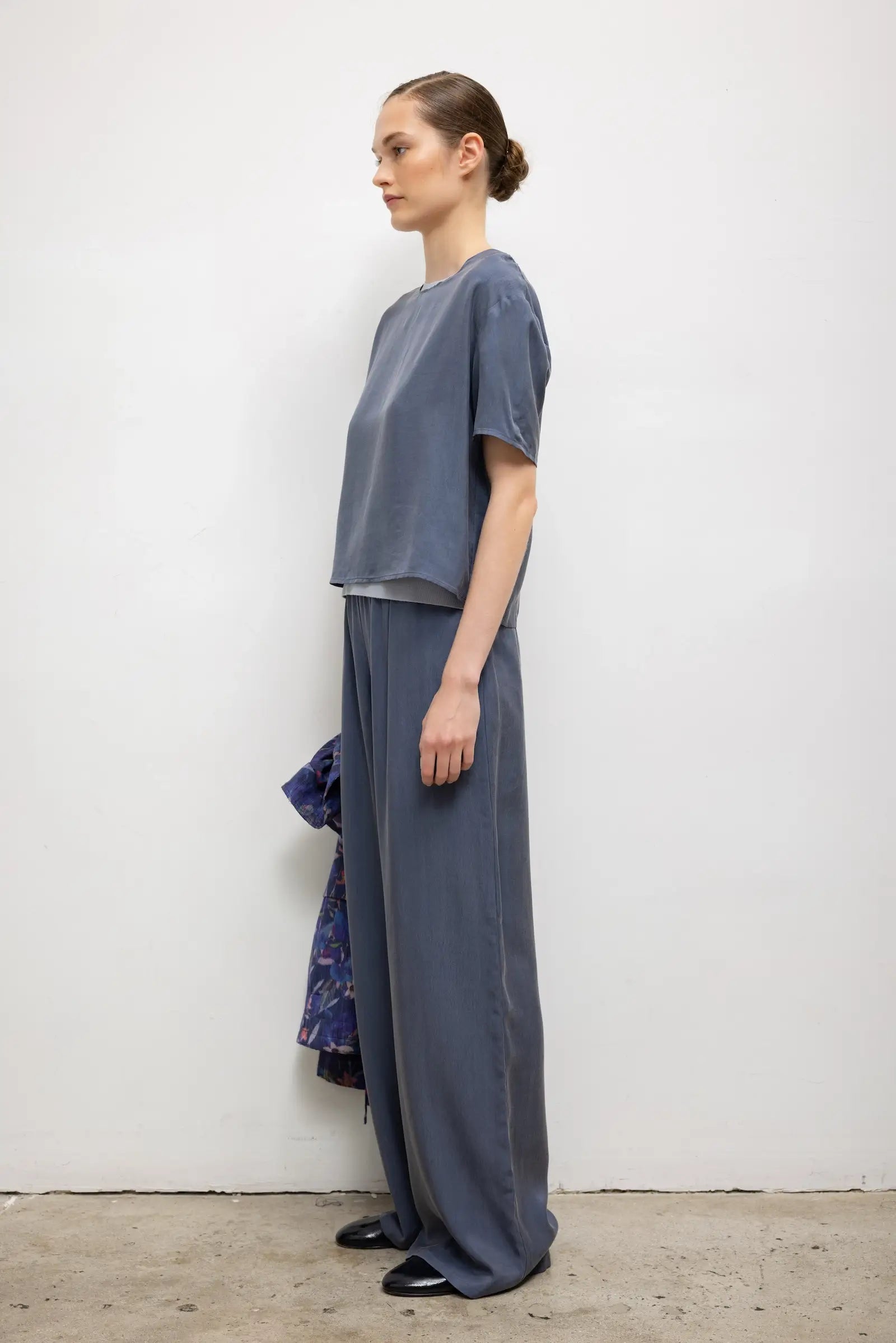 A woman stands in profile wearing a loose, short-sleeve gray-blue top and matching wide-leg pants, photographed against a plain off-white wall with a concrete floor.