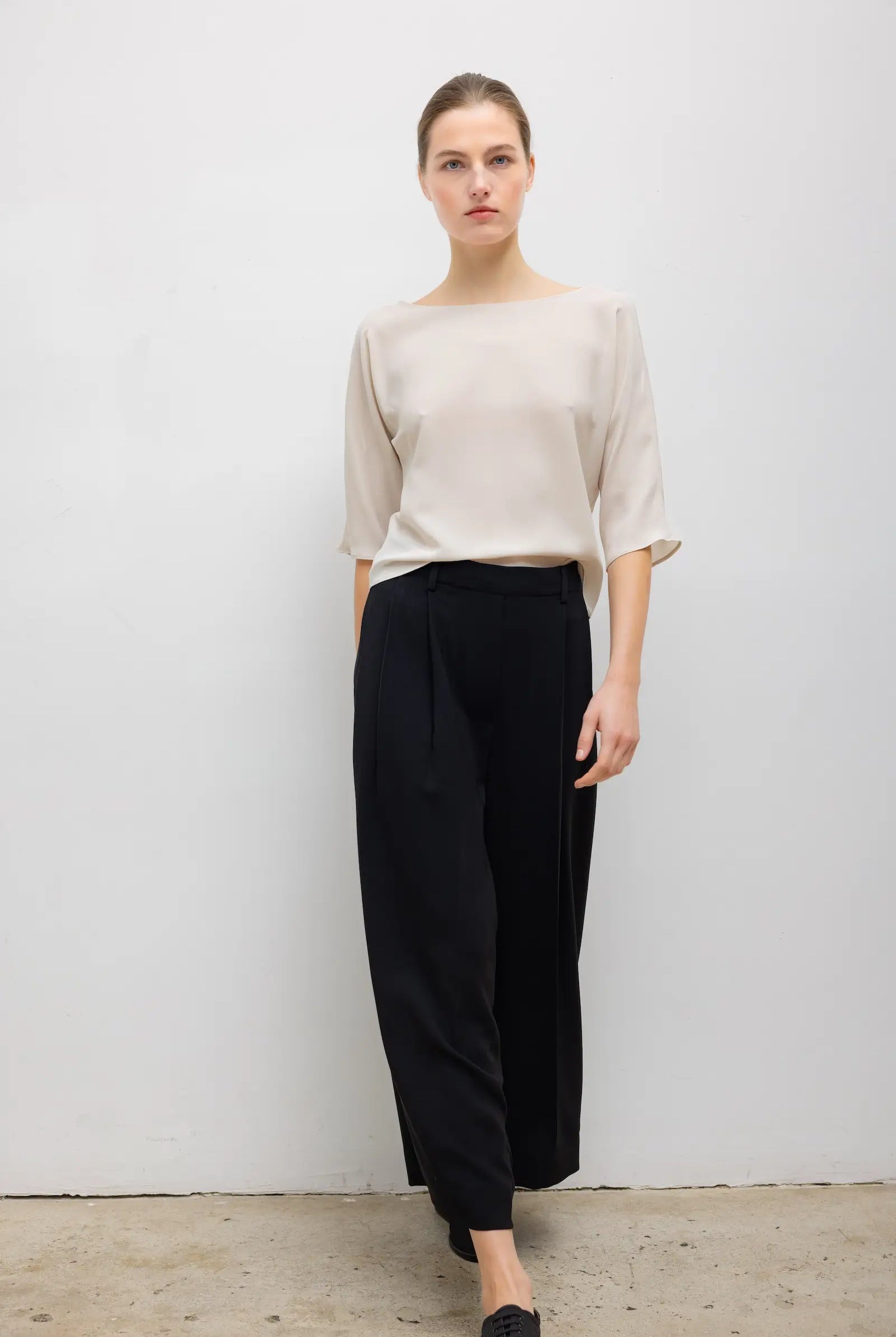 A woman modeling a light beige short-sleeve top and black wide-leg cropped trousers against a plain light gray wall.