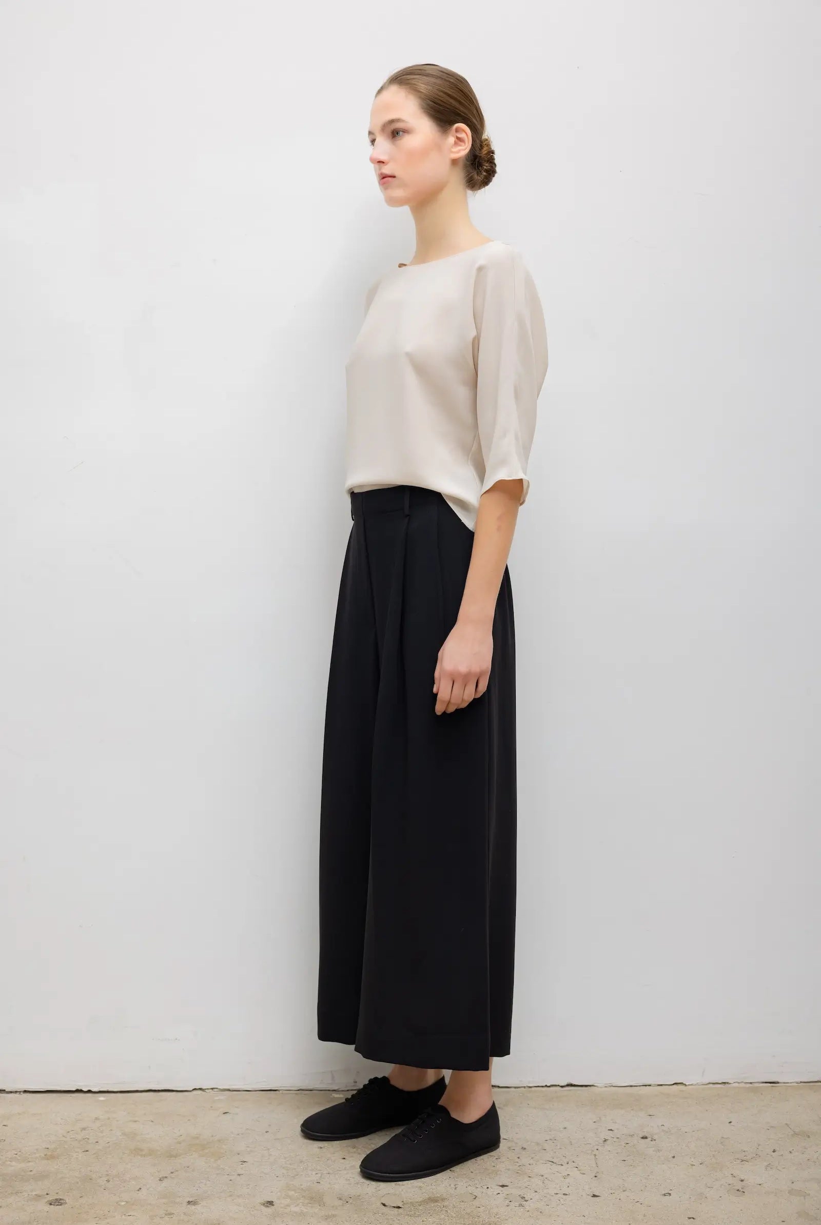 A woman stands in profile wearing a light beige short-sleeve top and black wide-leg cropped pants with black slip-on shoes, photographed against a plain light gray wall.