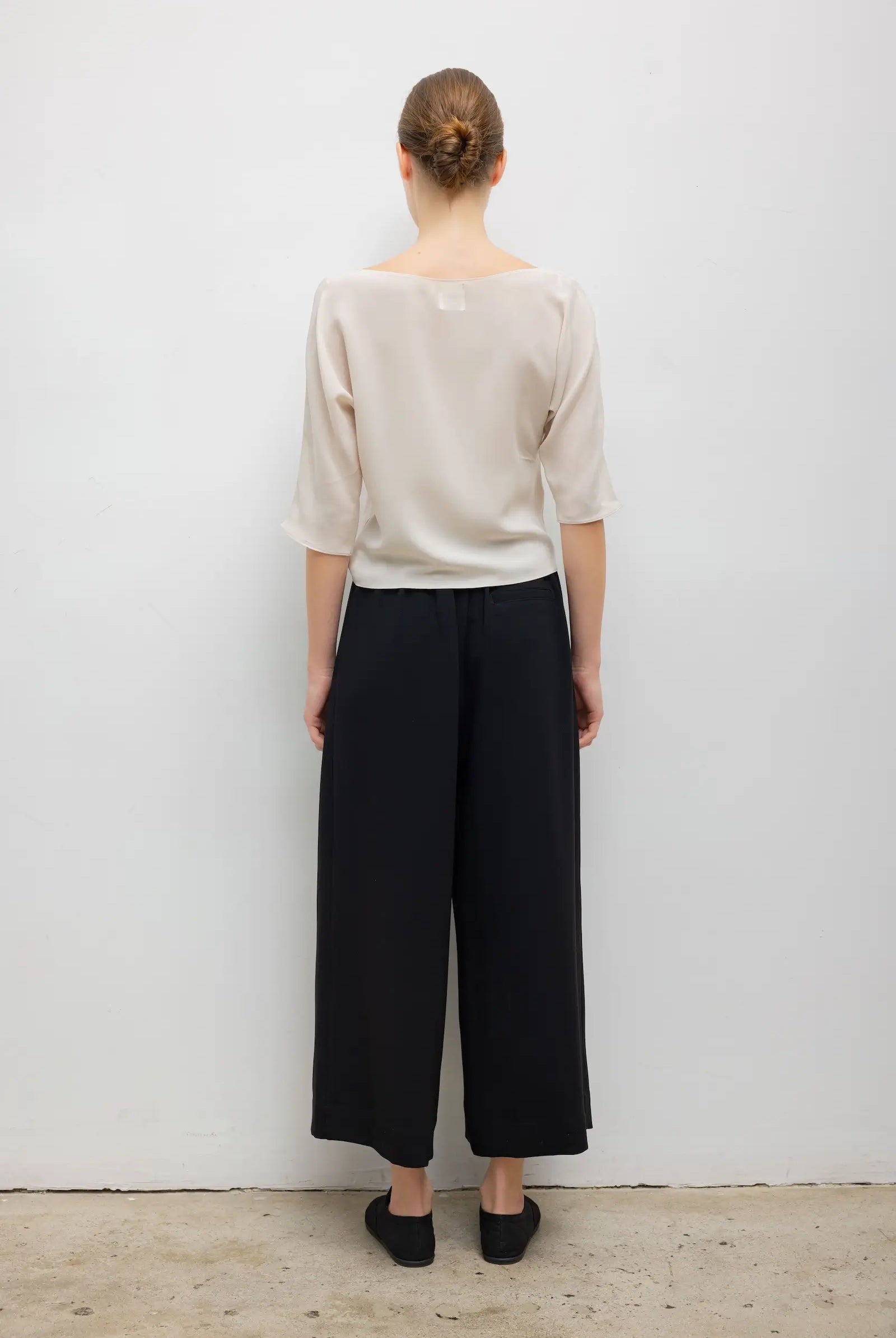 A woman shown from the back wearing a light beige short-sleeve top and black wide-leg cropped pants, standing against a plain white wall.