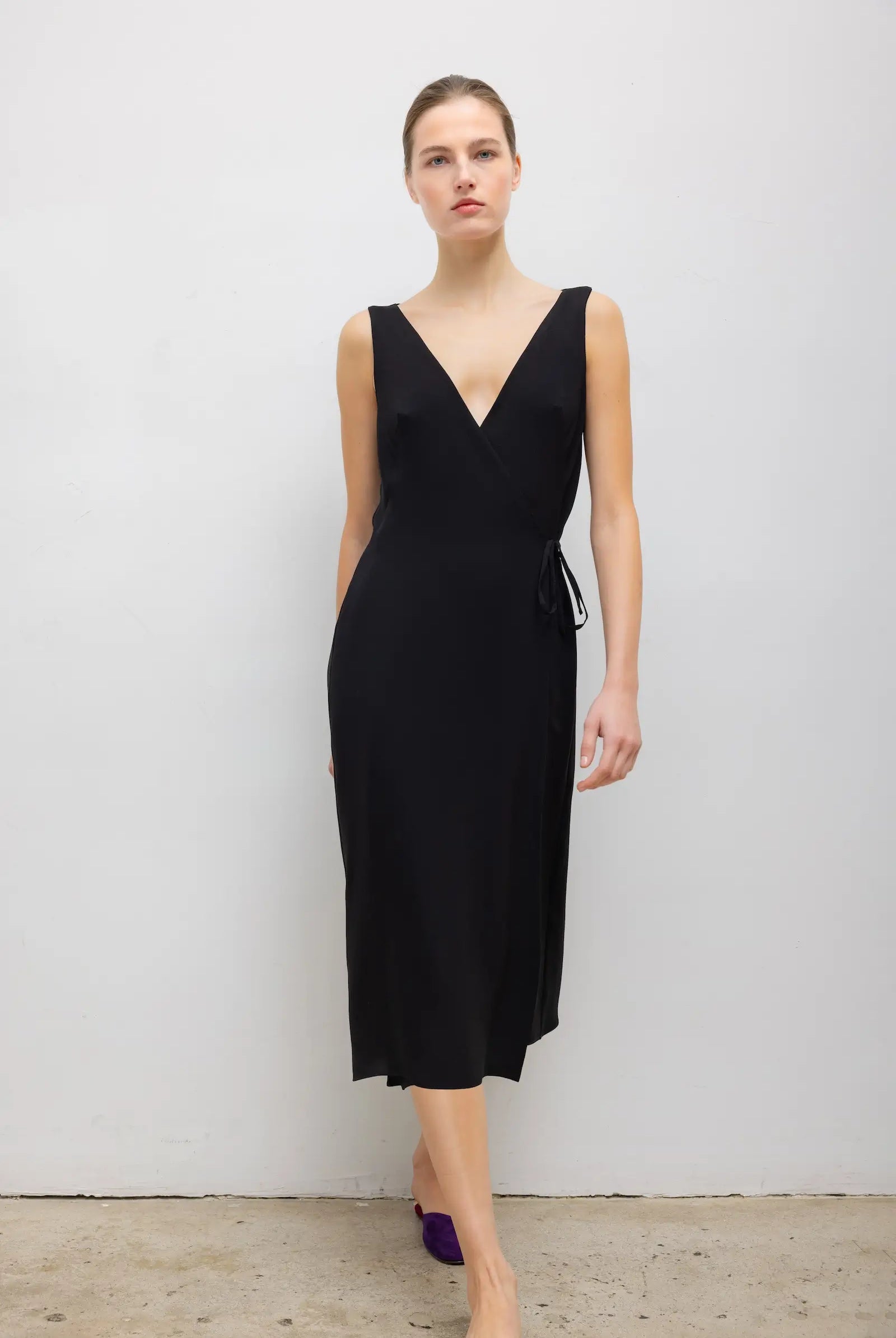 A woman modeling a sleeveless black midi dress with a wrap-style V-neck bodice and a side tie detail, standing against a plain light gray wall.
