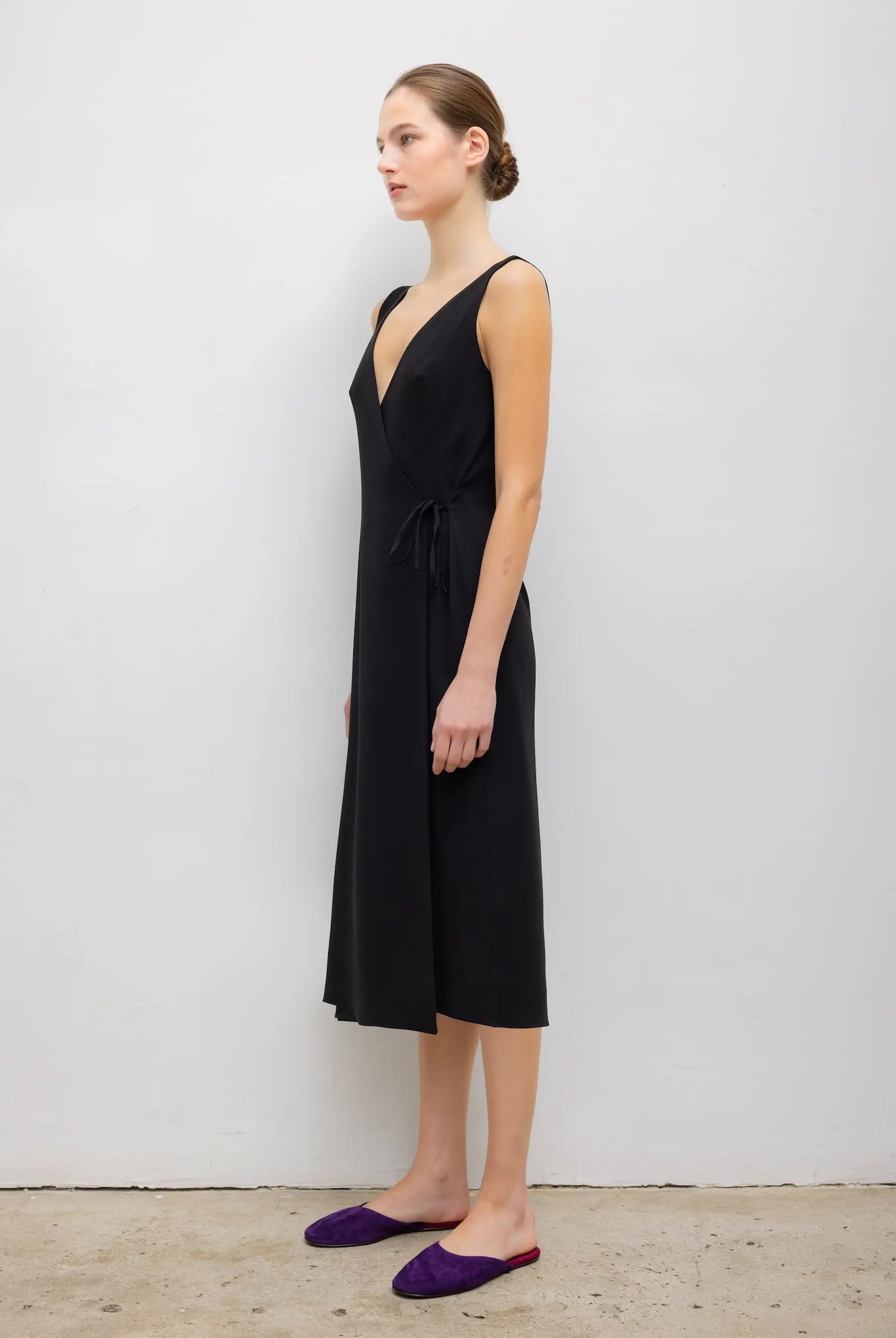 A woman stands in profile wearing a sleeveless black midi dress with a deep V-neck and a self-tie waist, paired with purple pointed flats, against a plain light gray wall.