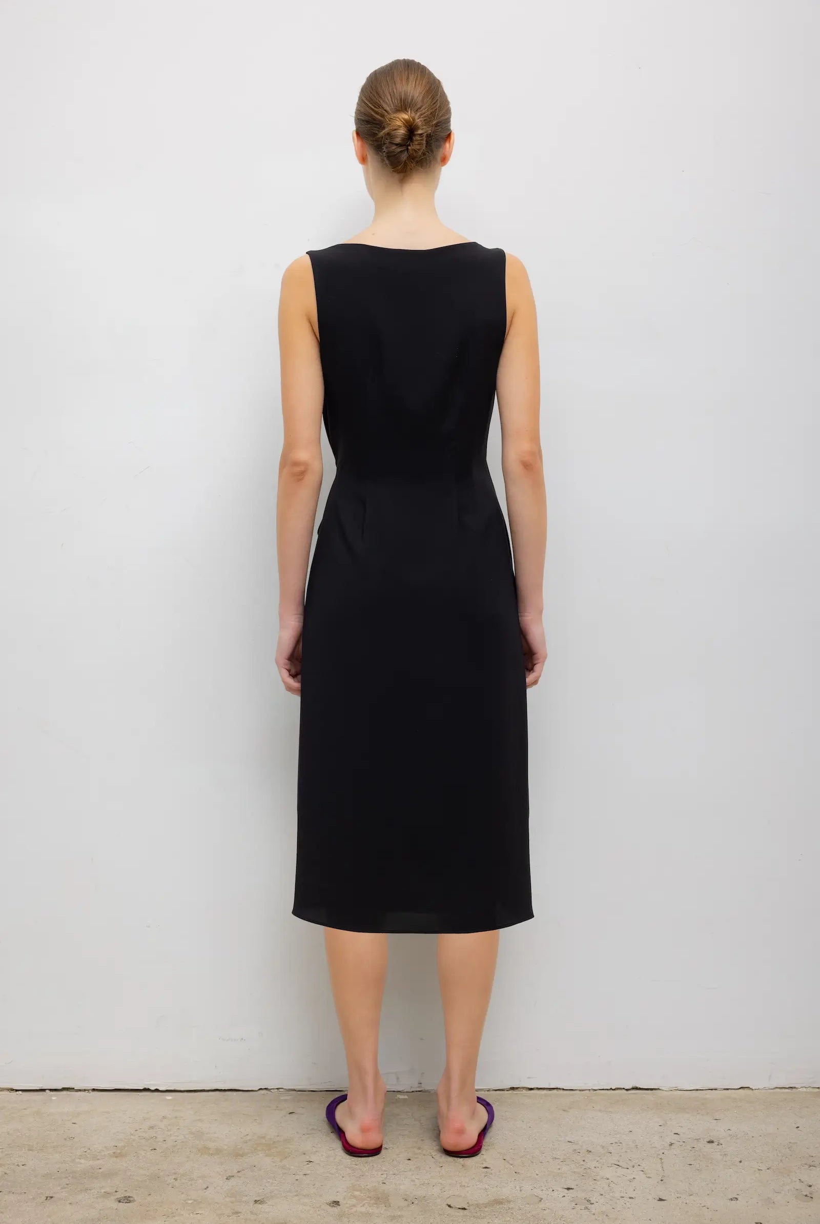 A woman viewed from the back wearing a sleeveless black knee-length dress, standing against a plain light gray wall on a concrete floor.