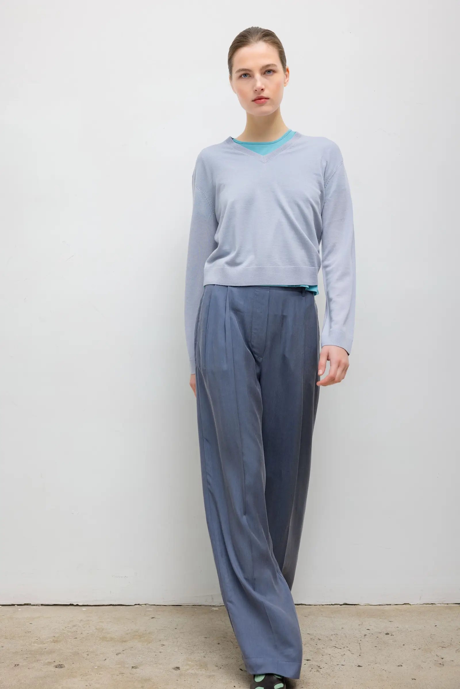 A woman modeling a light blue long-sleeve knit top and slate blue wide-leg trousers against a plain off-white wall.