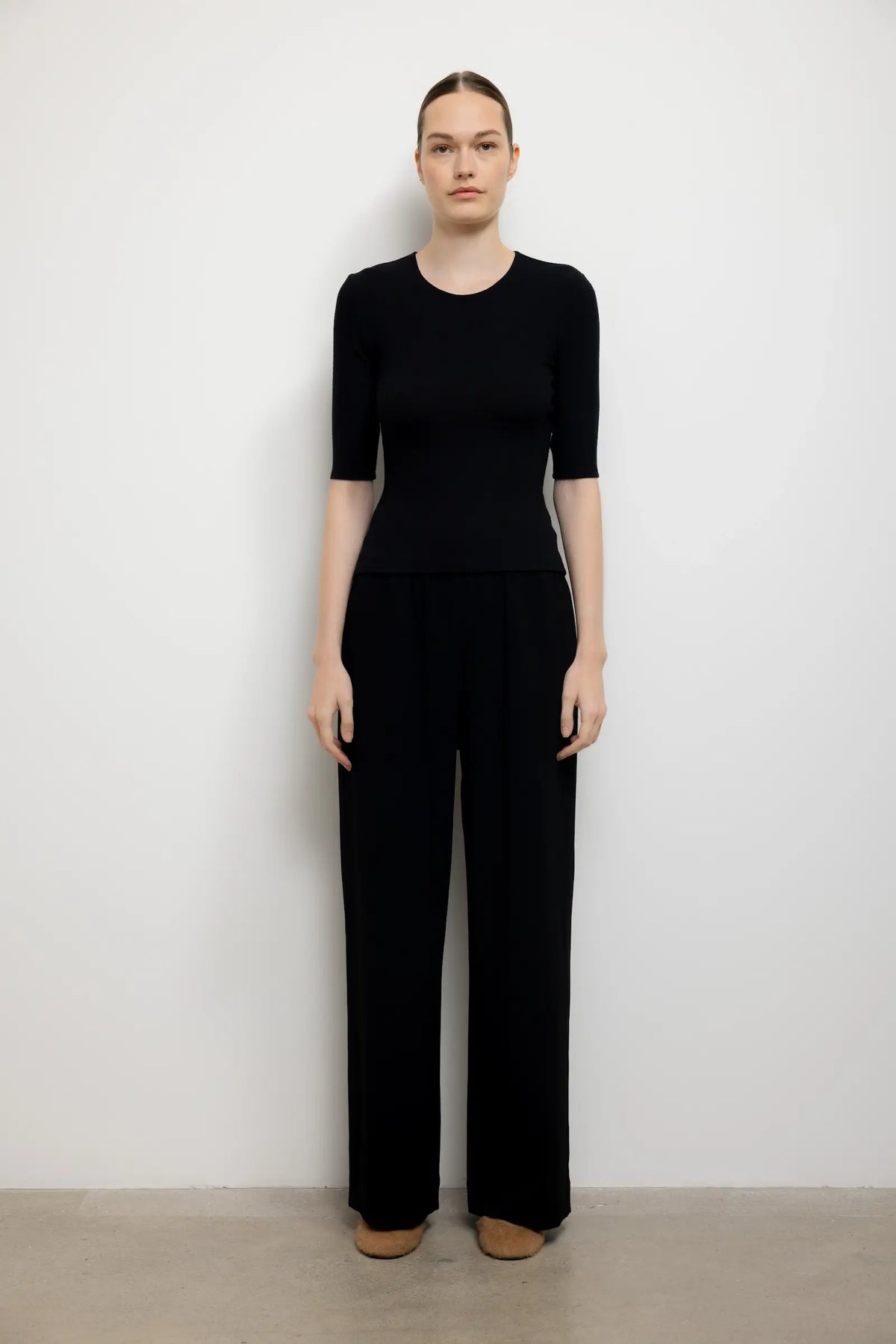 A model stands against a plain white wall wearing a black short-sleeve top and matching black wide-leg trousers, paired with tan shoes.