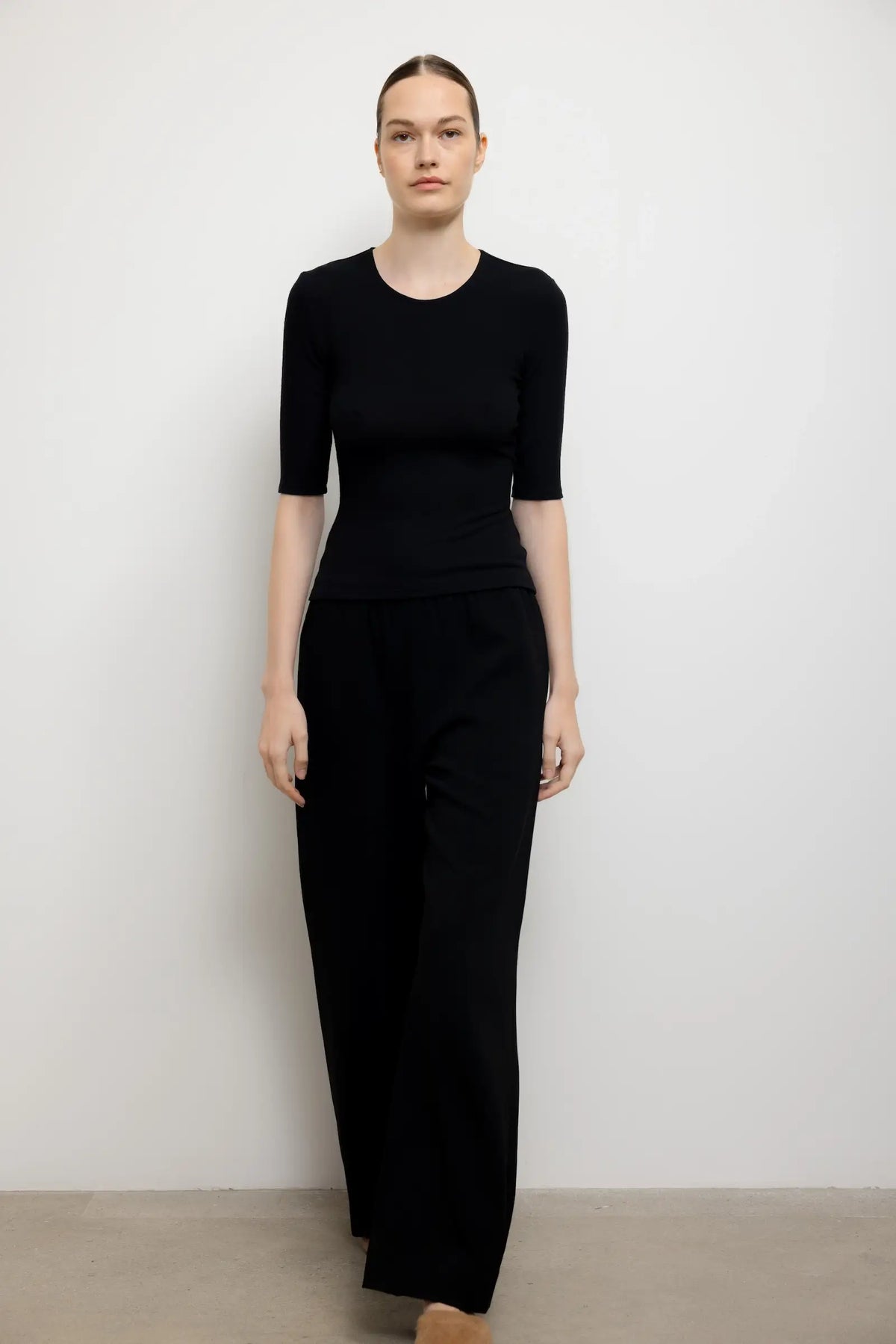 A woman modeling a black short-sleeve crewneck top and matching black wide-leg trousers against a plain light gray wall.