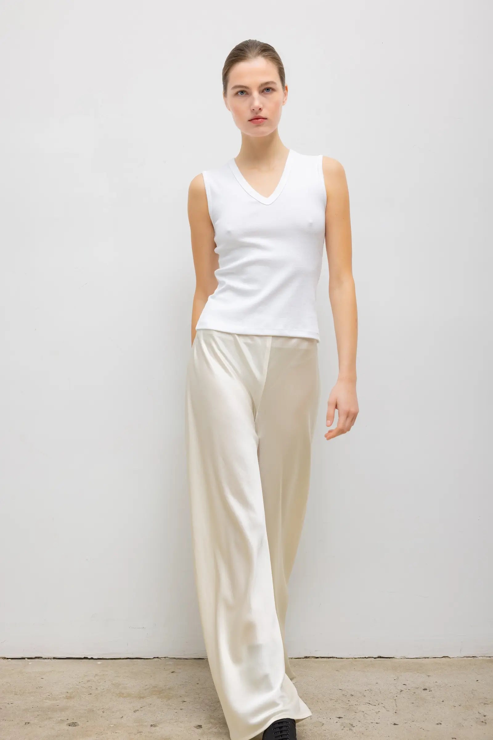 A woman modeling a sleeveless white V-neck top and wide-leg cream satin pants against a plain light gray wall.