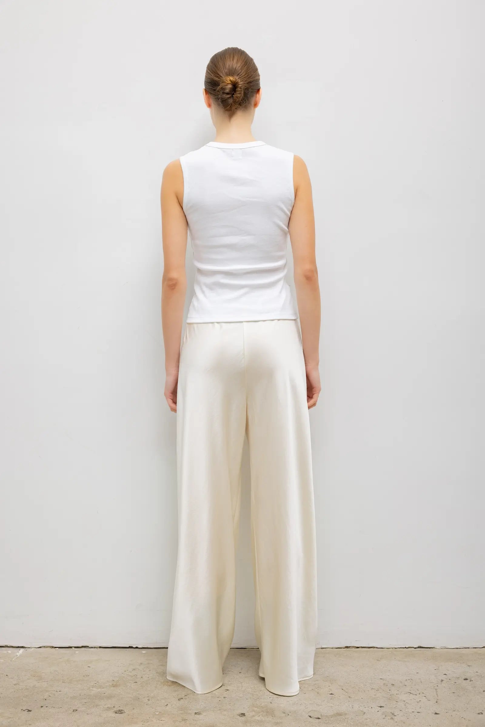 A woman viewed from behind wearing a white sleeveless top and cream wide-leg trousers, standing against a plain light gray wall.