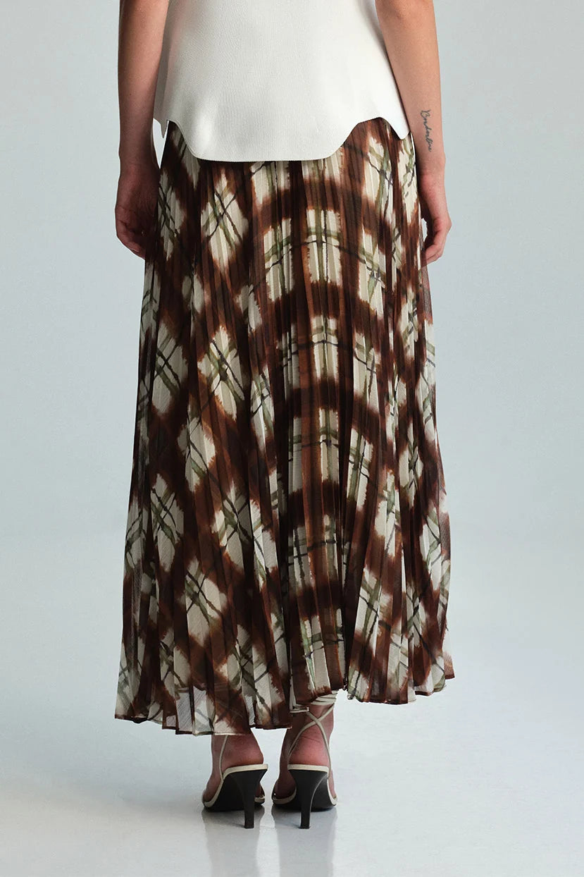 A woman shown from the back wearing a long pleated maxi skirt with a brown, beige, and black tie-dye pattern, paired with a white top and black high heels.