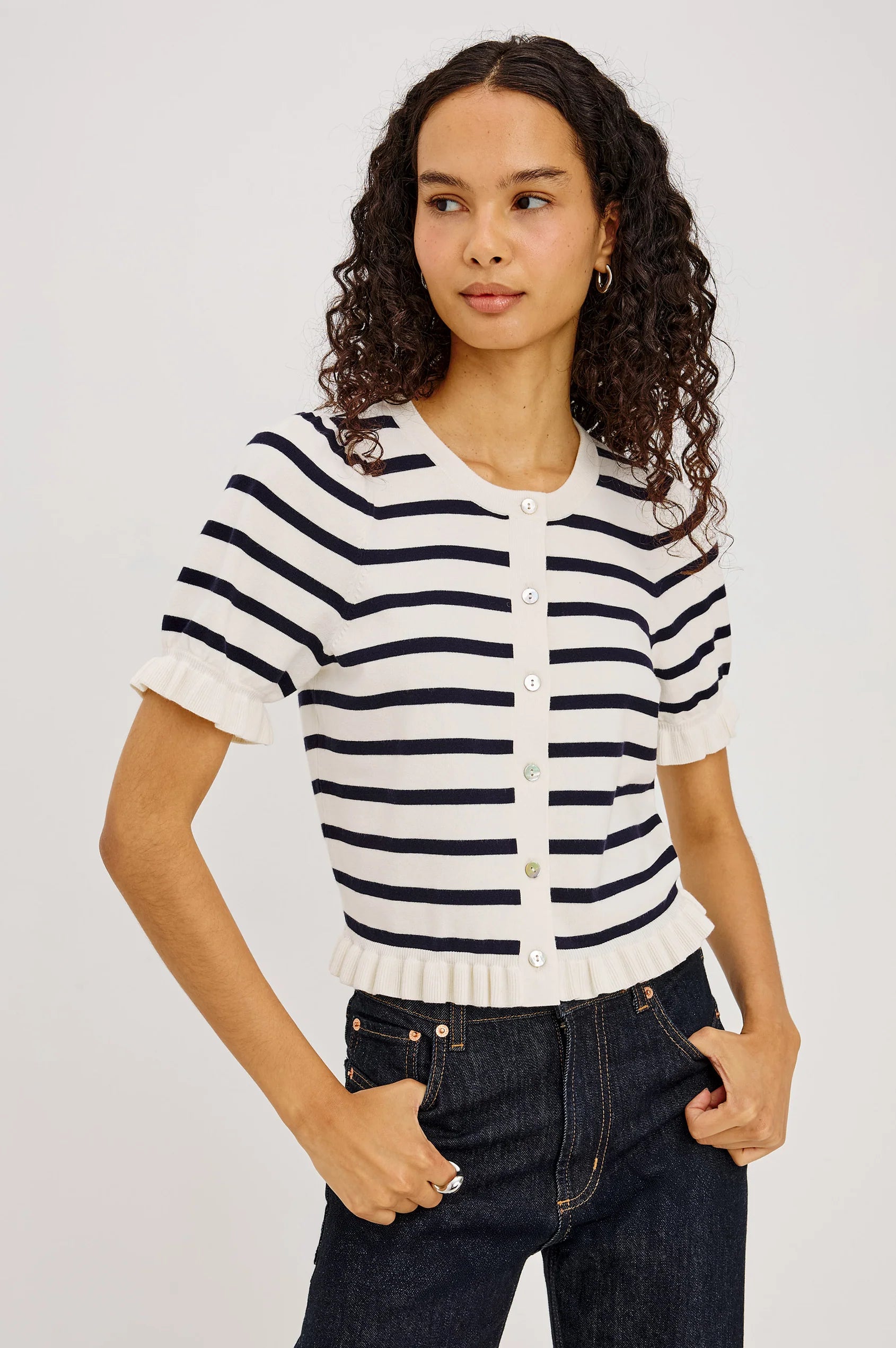 A woman with curly dark hair wearing a short-sleeve knit top with navy and white horizontal stripes, paired with dark blue jeans, against a light gray studio background.