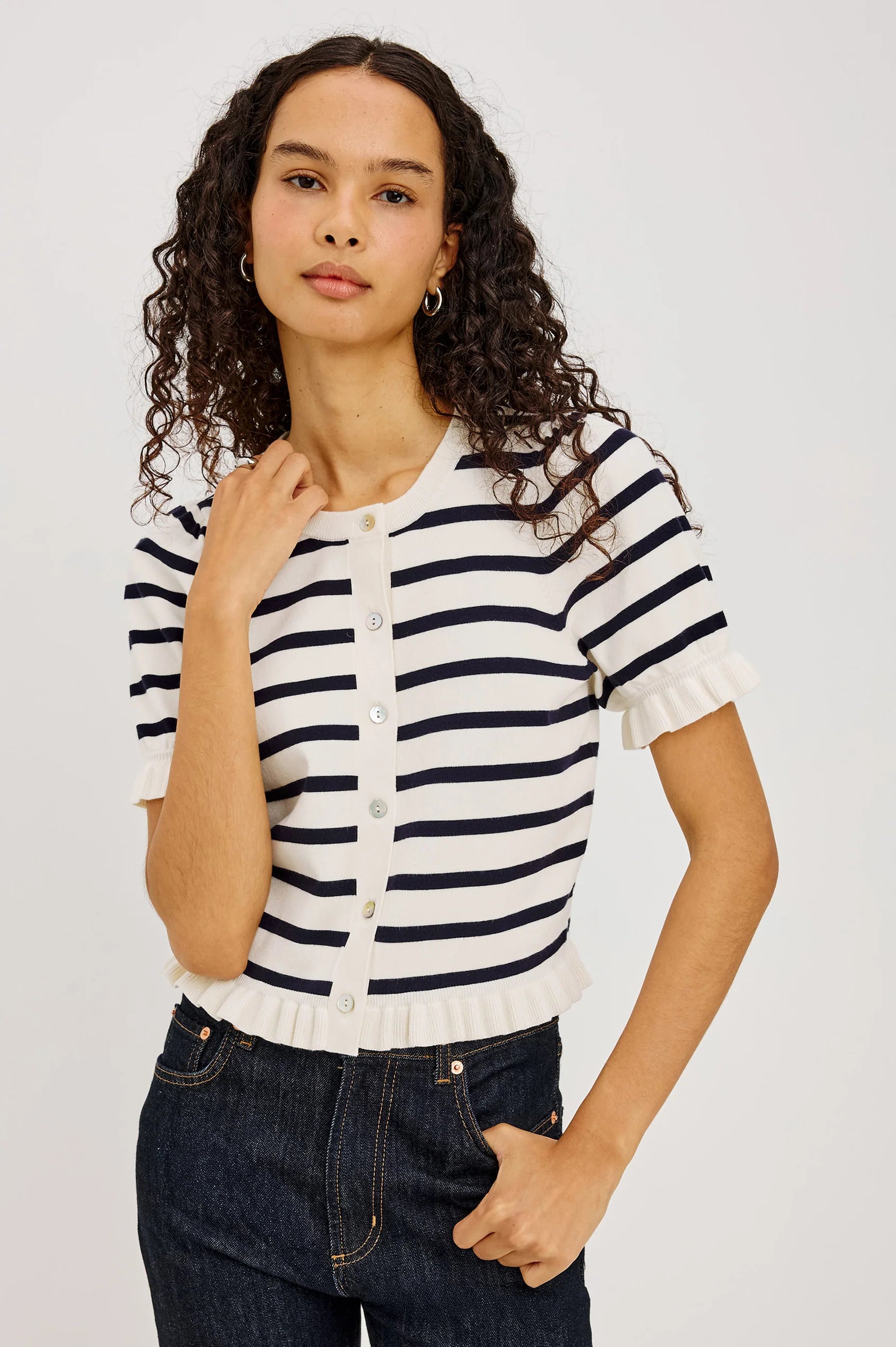 A woman with curly dark hair wearing a short-sleeve knit top with navy and white horizontal stripes, paired with dark blue jeans, against a light gray studio background.