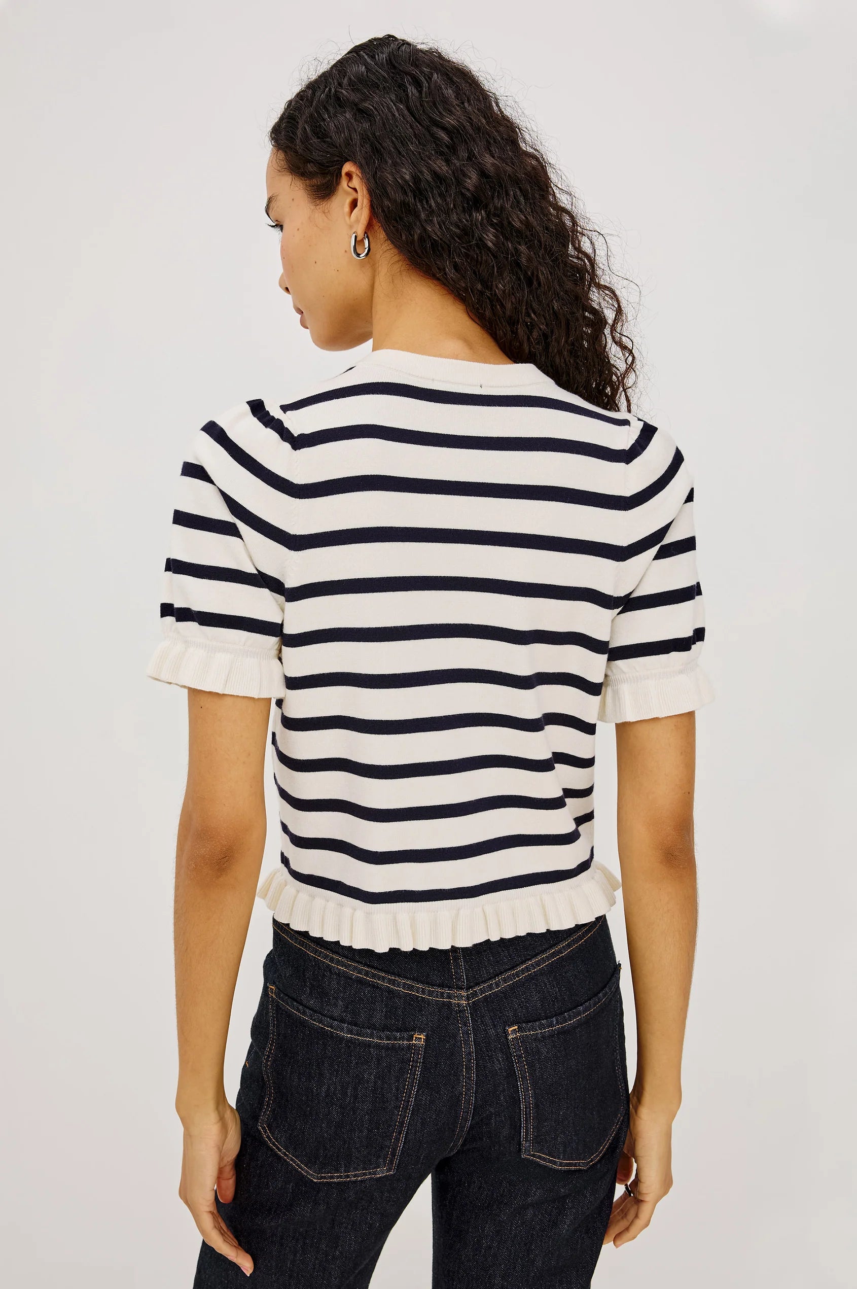 A woman shown from the back wearing a short-sleeve knit top with horizontal navy and off-white stripes, paired with dark blue jeans, against a light gray studio background.