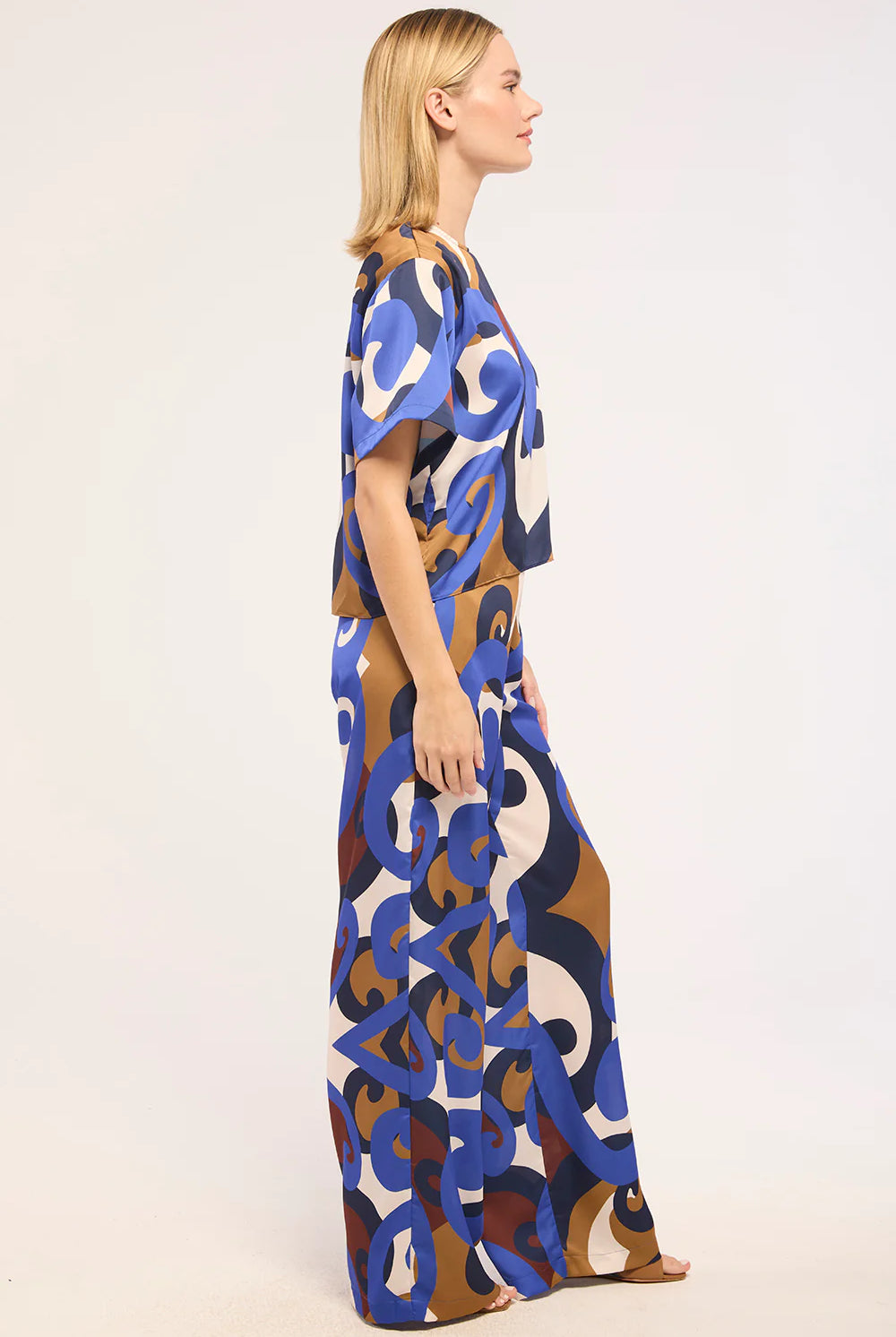 A woman modeling a short-sleeved maxi dress with an abstract, painterly pattern in blue, brown, beige, and white, shown from a side profile against a plain white studio background.
