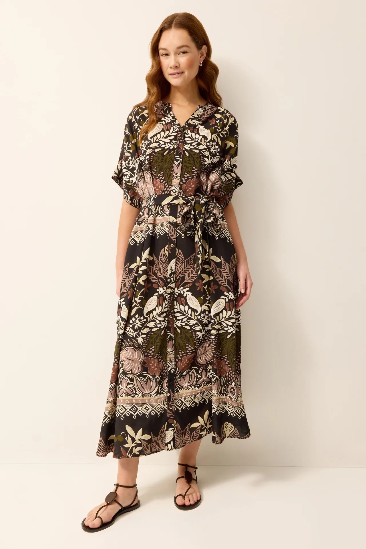 A woman modeling a short-sleeve midi dress with a botanical and tribal pattern in olive green, black, and beige, paired with black strappy sandals against a plain off-white background.