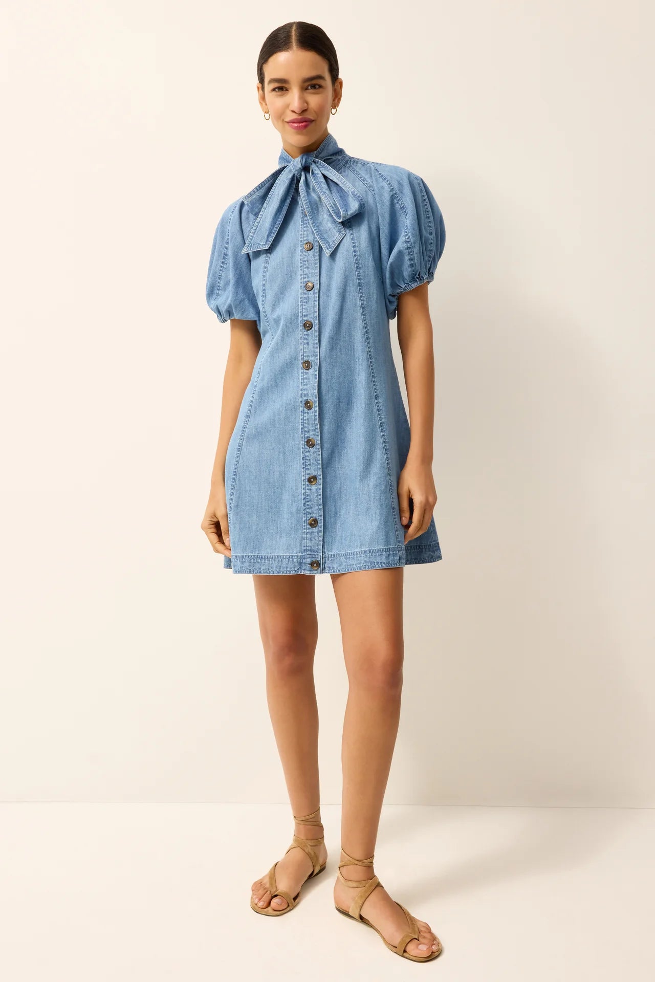 A model wearing a light blue denim mini dress with puff sleeves, a tie-neck bow, and a button-down front, paired with beige strappy sandals against a plain cream studio background.