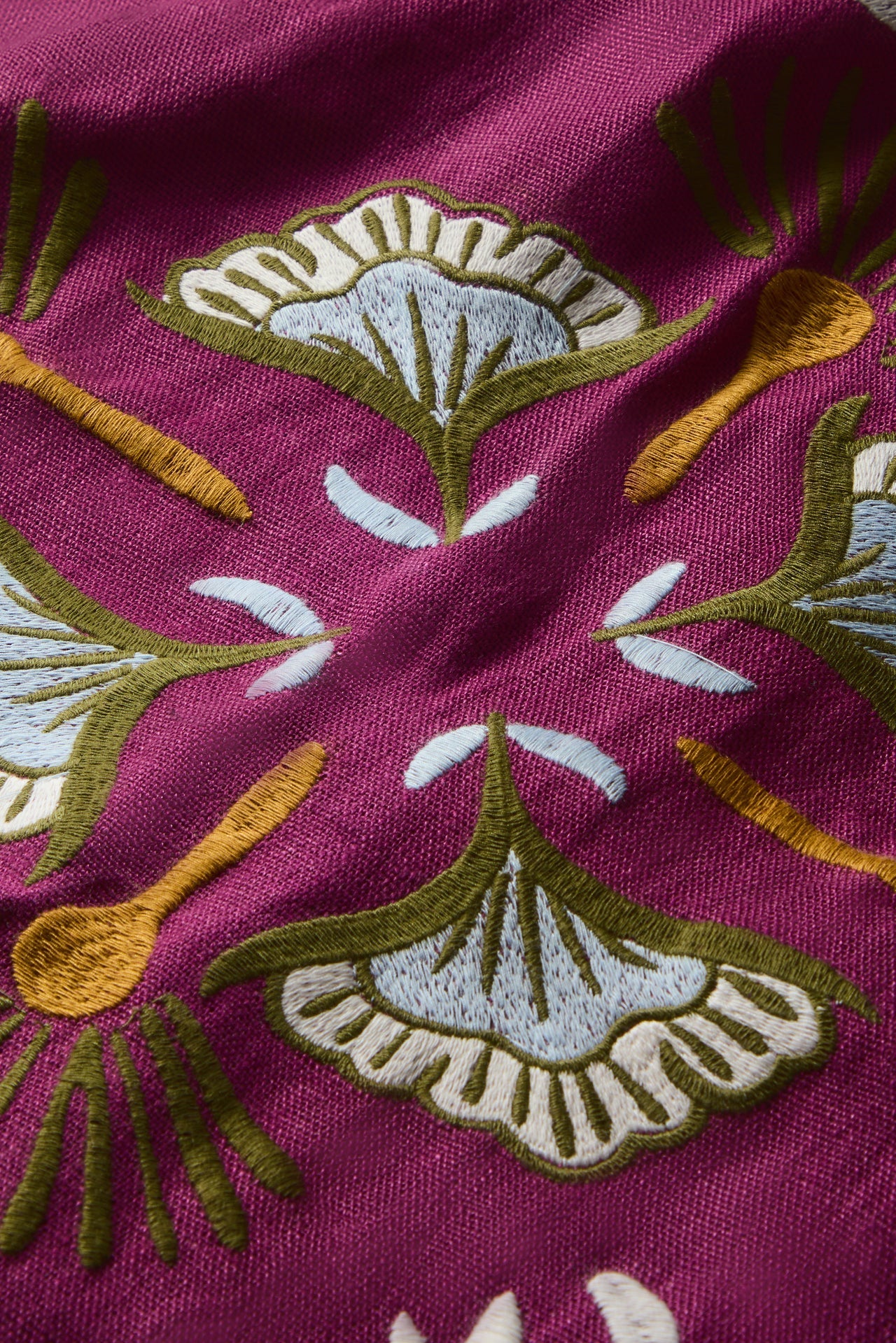 A close-up of a magenta fabric with embroidered botanical motifs in olive green, cream, and mustard yellow.