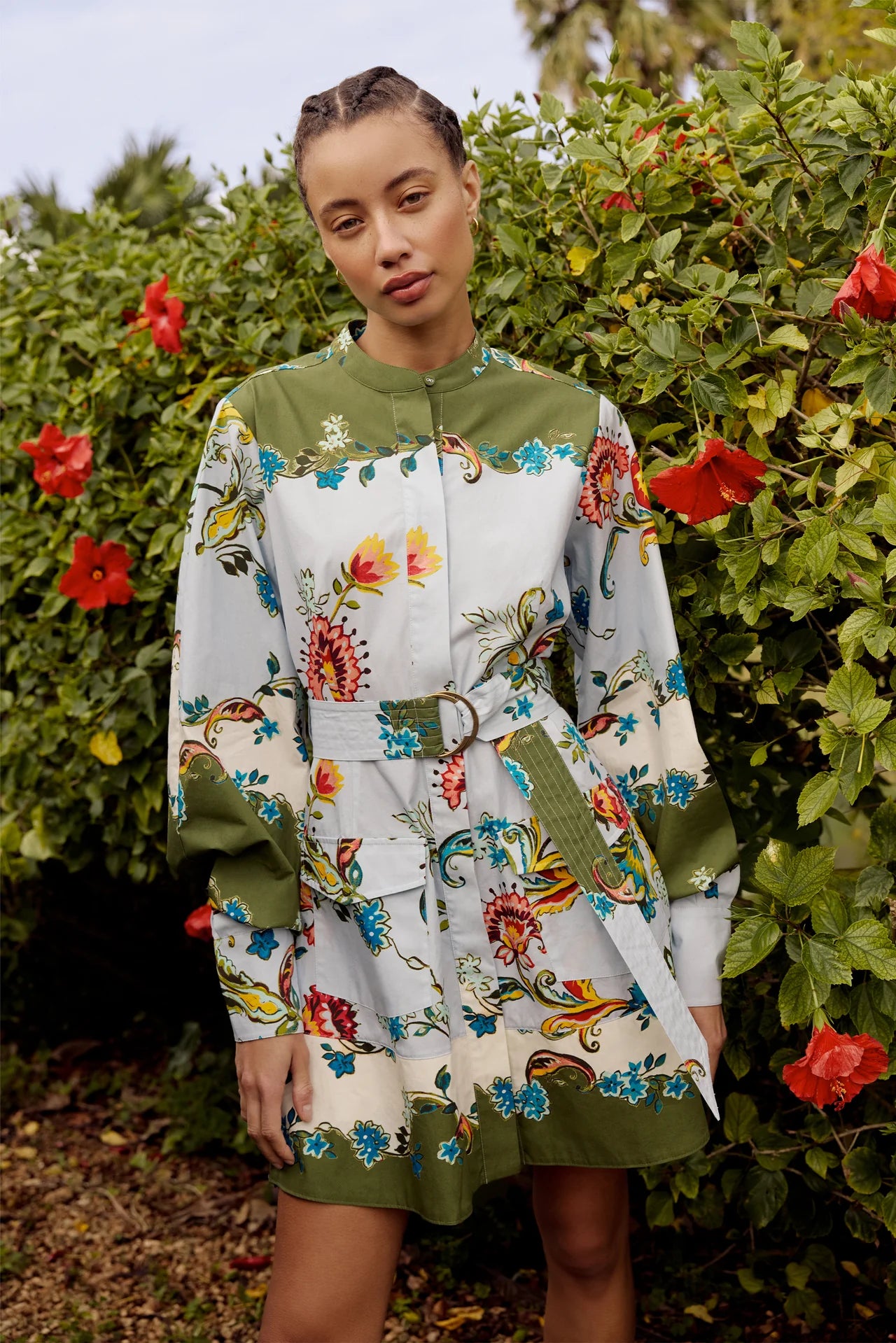 Woman wearing a floral dress with green accents standing in front of greenery with red flowers.