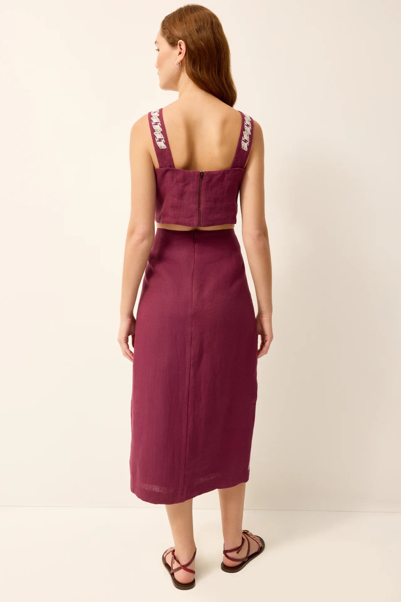 A woman viewed from the back wearing a sleeveless deep plum midi dress with a structured square neckline, a matching cropped top, and embroidered shoulder straps, paired with brown strappy sandals against a plain off-white studio background.