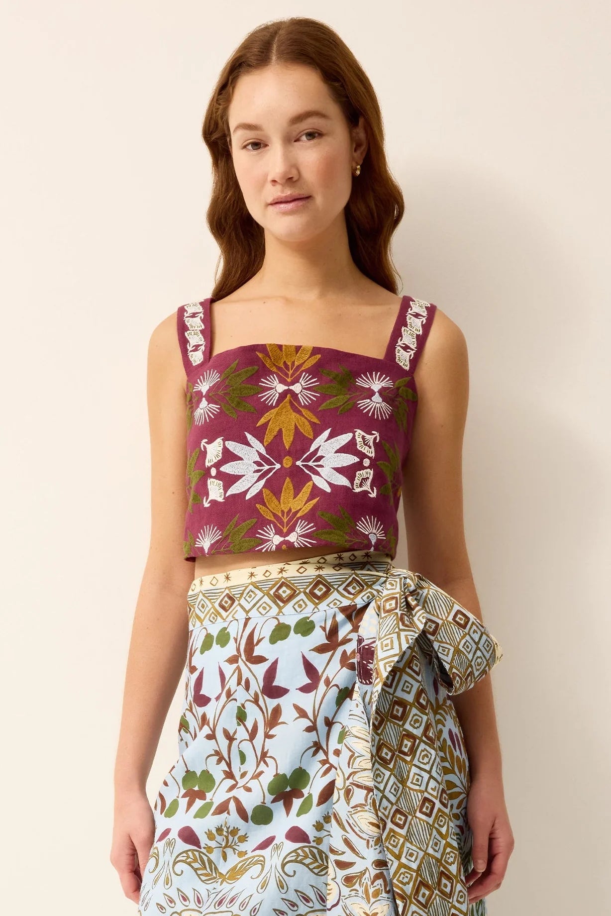 A woman modeling a sleeveless top and matching patterned skirt set against a plain beige studio background. The top has a deep magenta-burgundy base with white, olive green, and mustard yellow floral and geometric motifs.