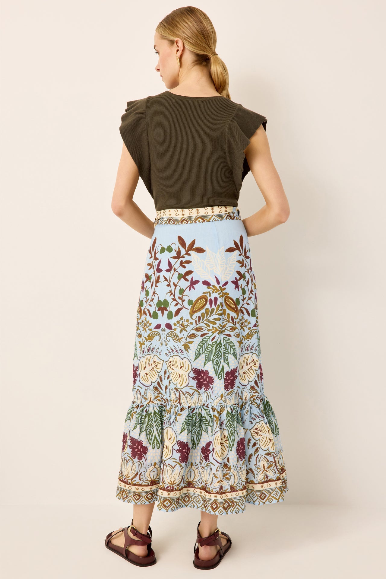 A woman shown from the back wearing a dark olive green short-sleeve top and a long light blue tiered skirt with a detailed floral and bird print, paired with brown sandals, against a solid beige background.