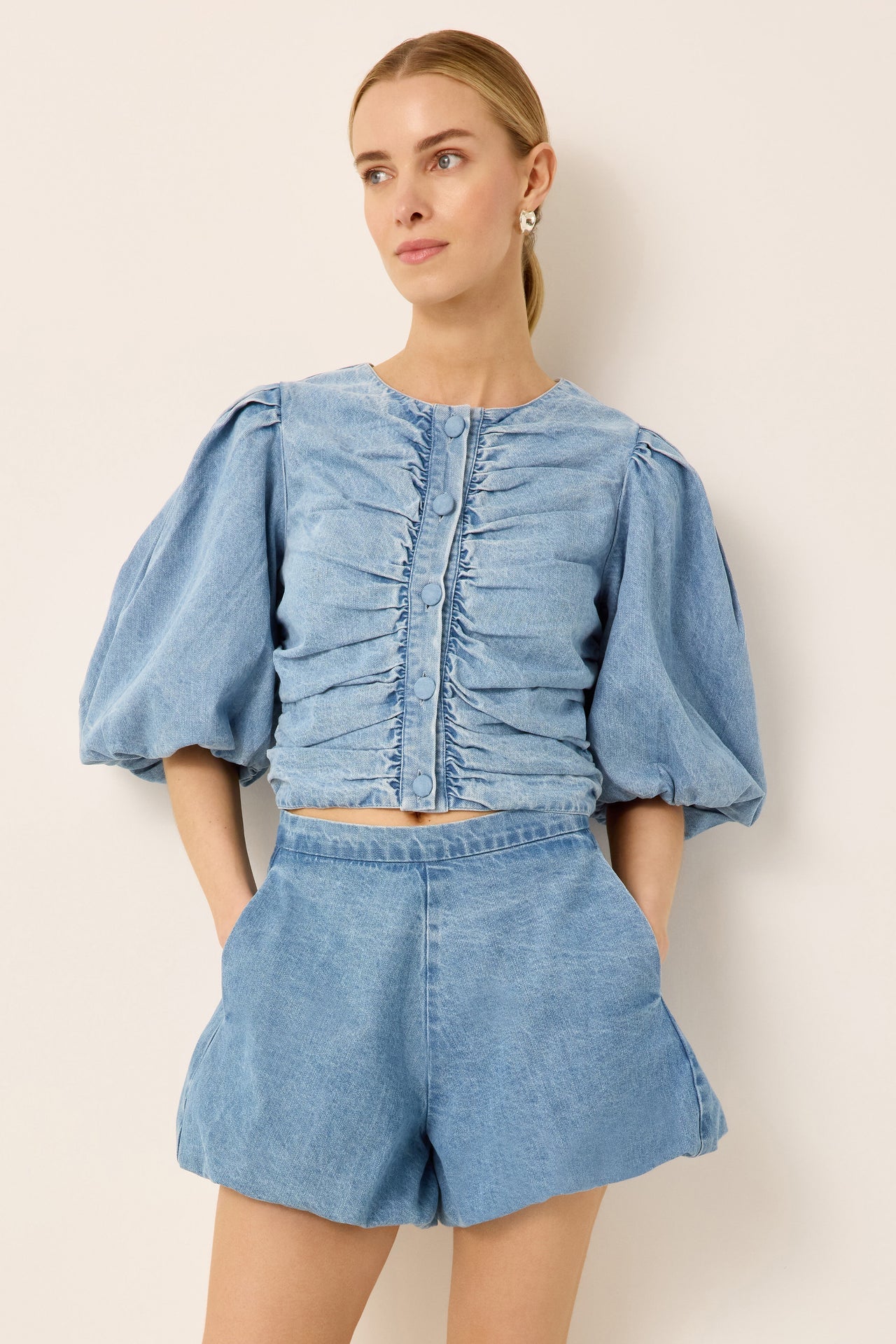 A woman modeling a light blue denim two-piece set consisting of a cropped top with dramatic puff sleeves and front pleating, paired with matching high-waisted shorts, against a plain beige background.