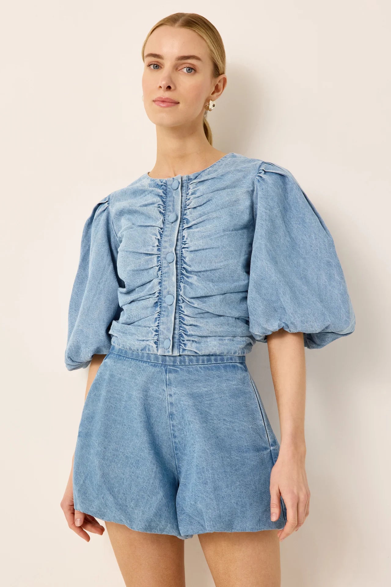 A woman modeling a light blue denim romper with a ruched front and puffed short sleeves, photographed against a plain light beige wall.