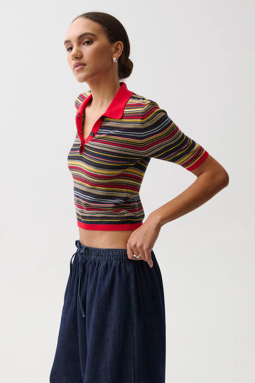 A woman modeling a short-sleeve cropped knit polo shirt with horizontal multicolor stripes and a red collar, paired with dark blue wide-leg pants, against a light gray studio background.