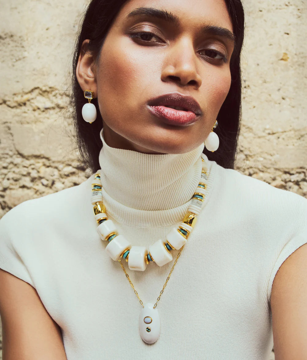 Woman wearing a white turtleneck and a long necklace with large beads against a stone wall.