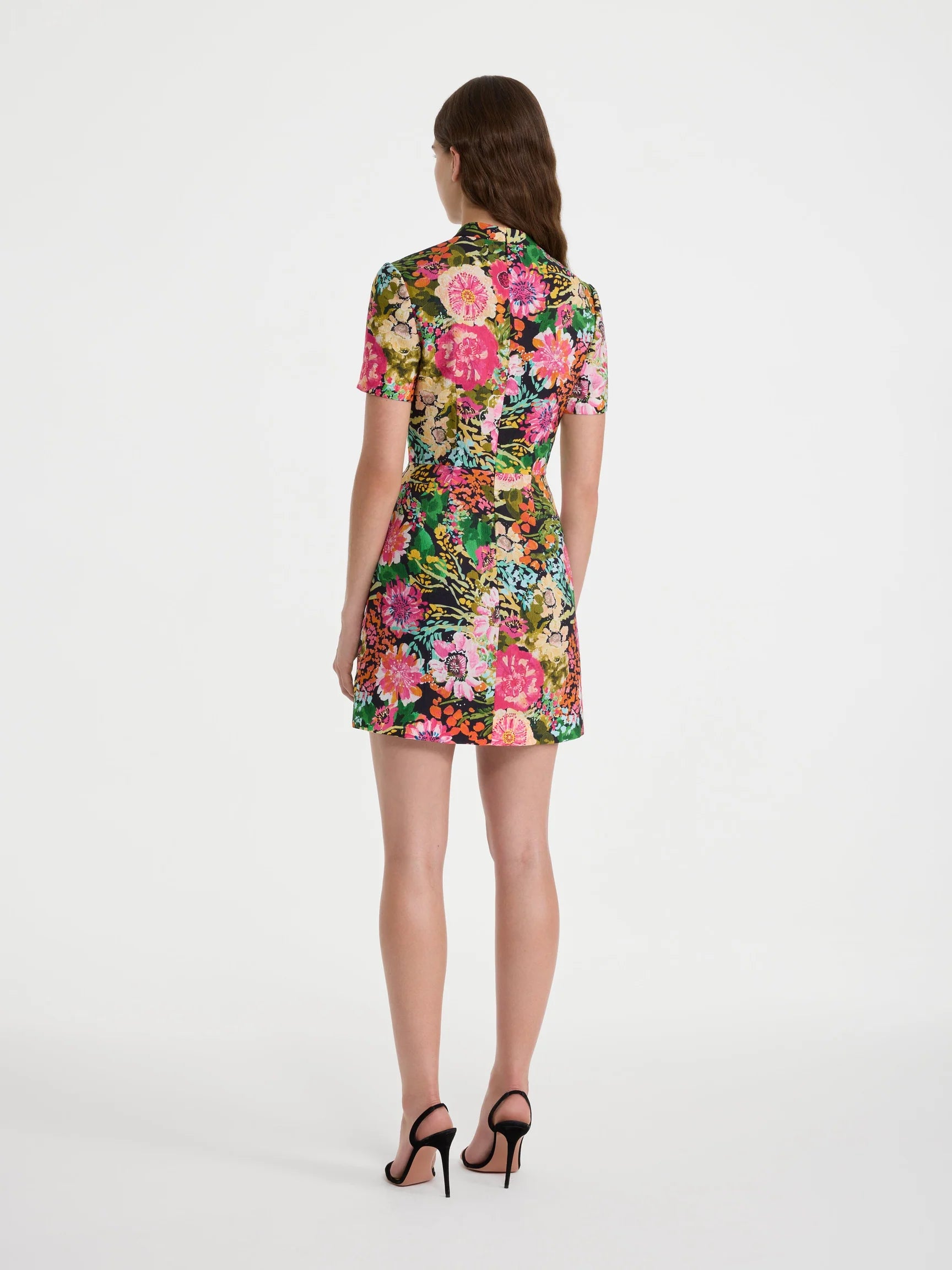 A woman viewed from the back wearing a short-sleeve collared mini dress with a dense multicolor floral print, paired with black high-heeled sandals, against a plain white studio background.