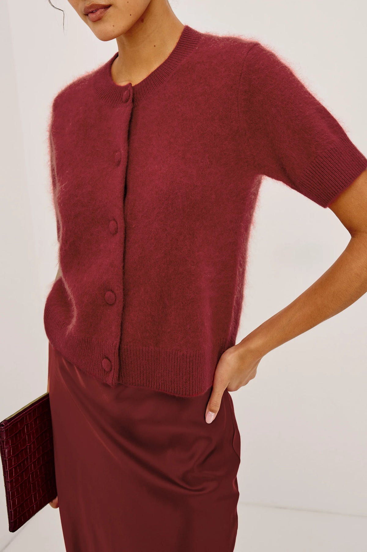 A close-up of a model wearing a short-sleeve fuzzy knit cardigan and matching satin skirt in a deep burgundy red, holding a matching clutch.